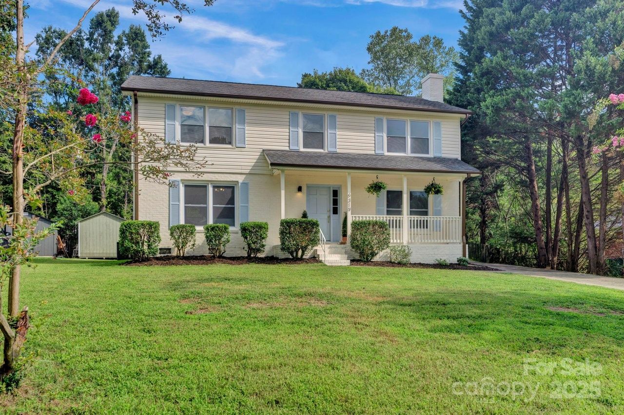 Two-story beige house with gray shutters, front porch with hanging plants, green lawn, bushes, trees, and blue sky. A small shed is visible on the left side.