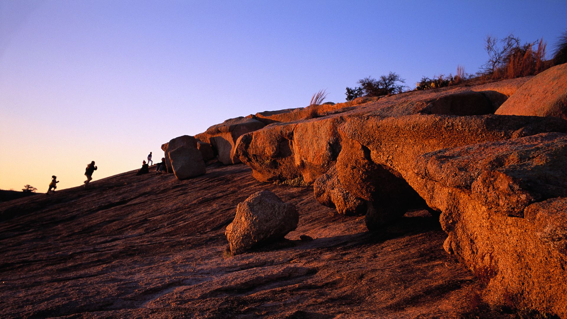 Hikers climb on a large rock that covers the ground at sunset.