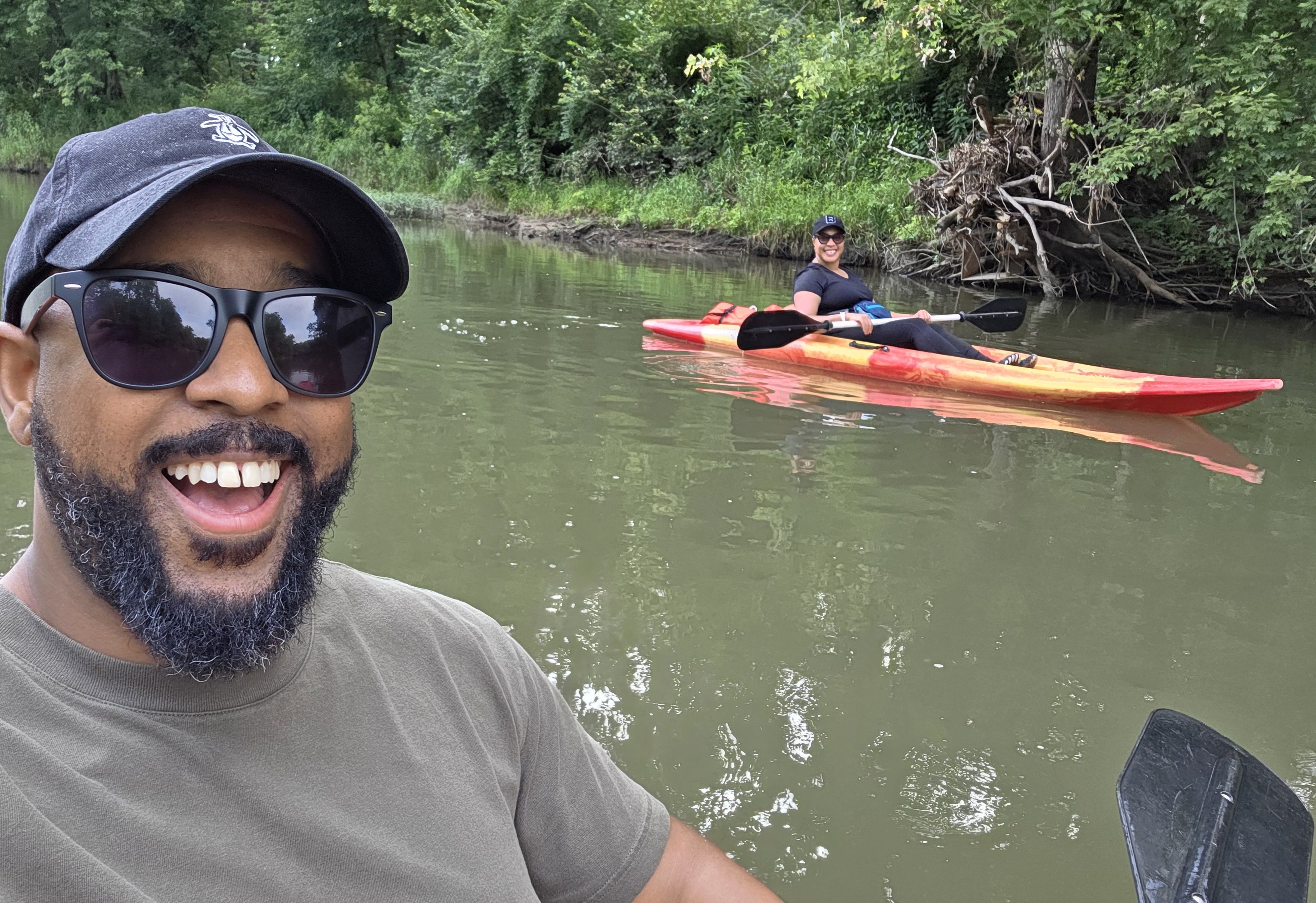 Smiling man wearing sunglasses and a black cap takes selfie while a woman in a red and yellow kayak paddles on a calm river surrounded by green trees.