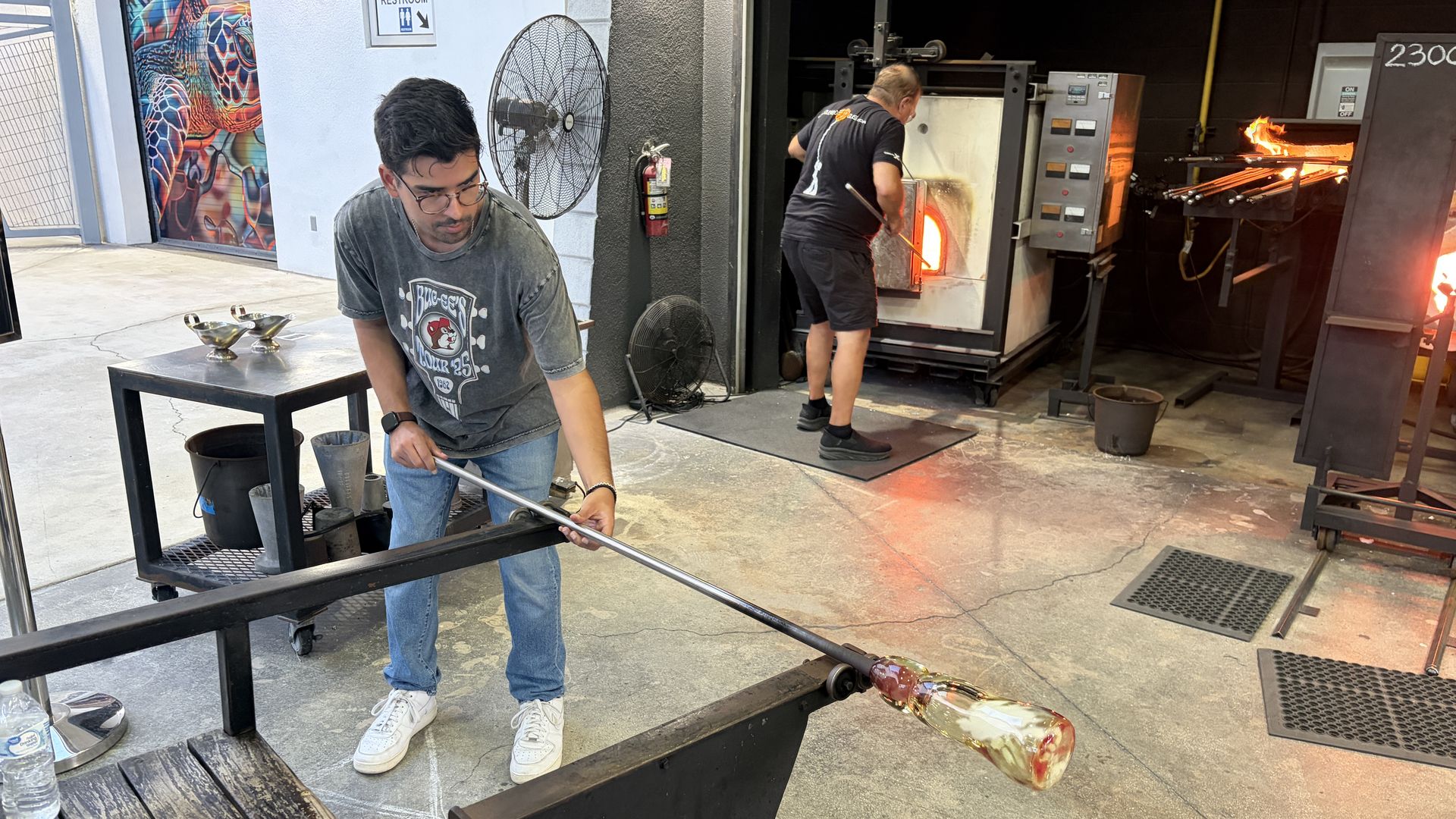 Two glassblowers work in a studio by a glowing furnace; one in front with glasses looks down, another feeds molten glass on a long rod. Nearby: fans, tools, water bottle, mural door.