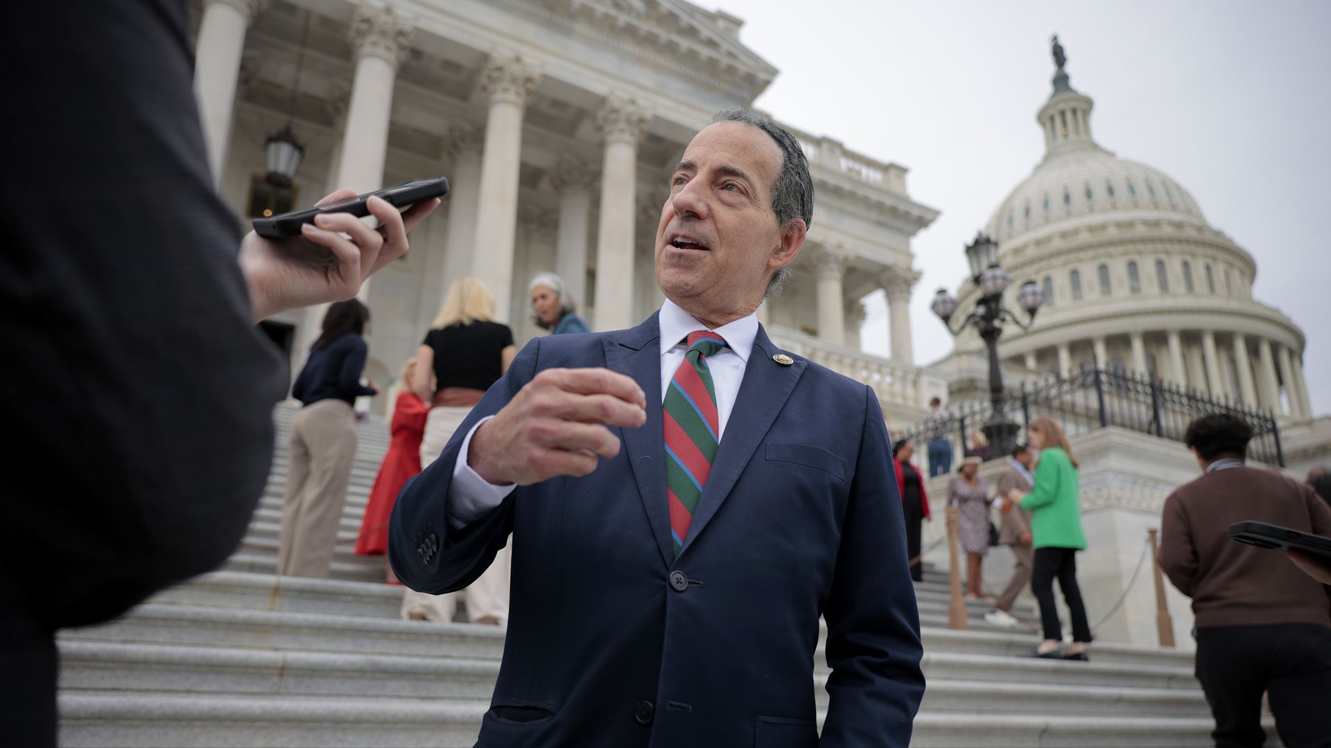 Rep Jamie Raskin, in a blue suit with a red and green striped ti and the Capitol dome in the background, answers a reporter's question.