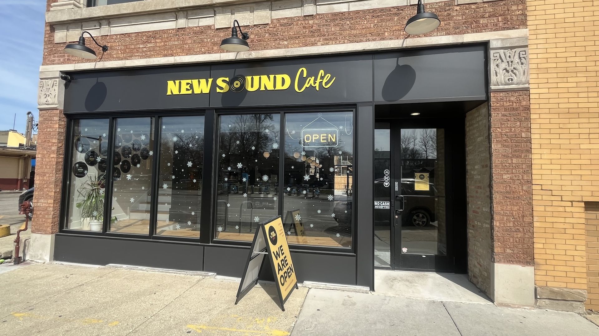 Front of New Sound Cafe storefront: black facade with yellow lettering, large windows with snowflake decals, neon OPEN sign, and a yellow WE ARE OPEN A-frame on the sidewalk.