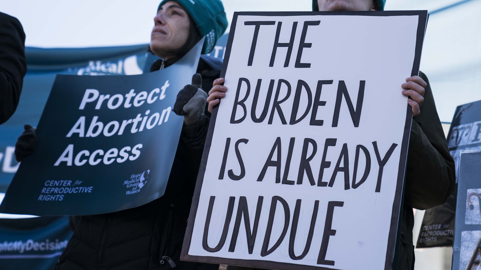 Photo of a person holding a sign that says "The burden is already undue" and another person holding a sign that says "Protect abortion access"