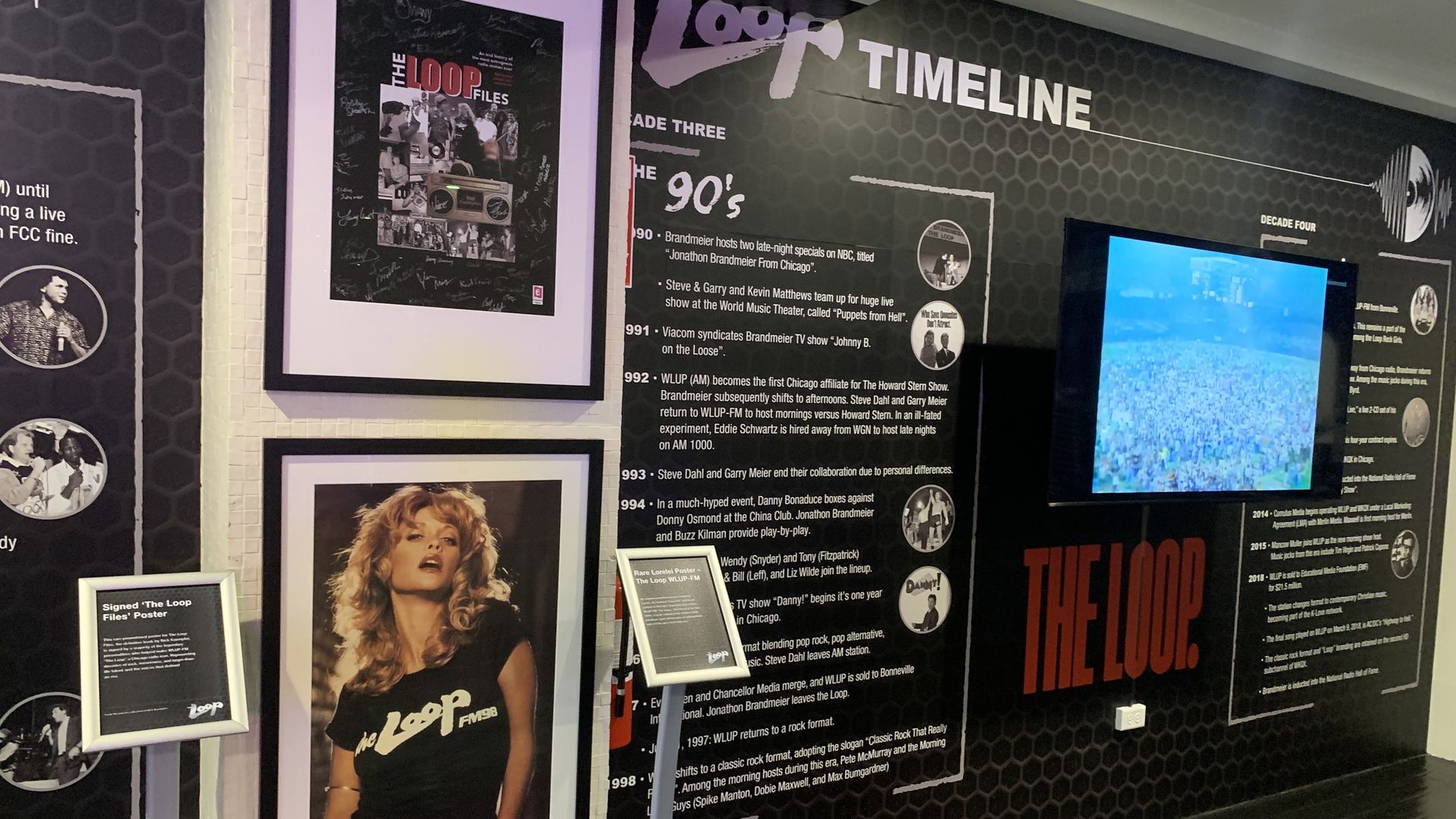 Wall exhibit featuring The Loop timeline with black honeycomb background, framed signed poster and photo of woman in a black "The Loop FM 98" shirt, and a TV screen showing a crowd.