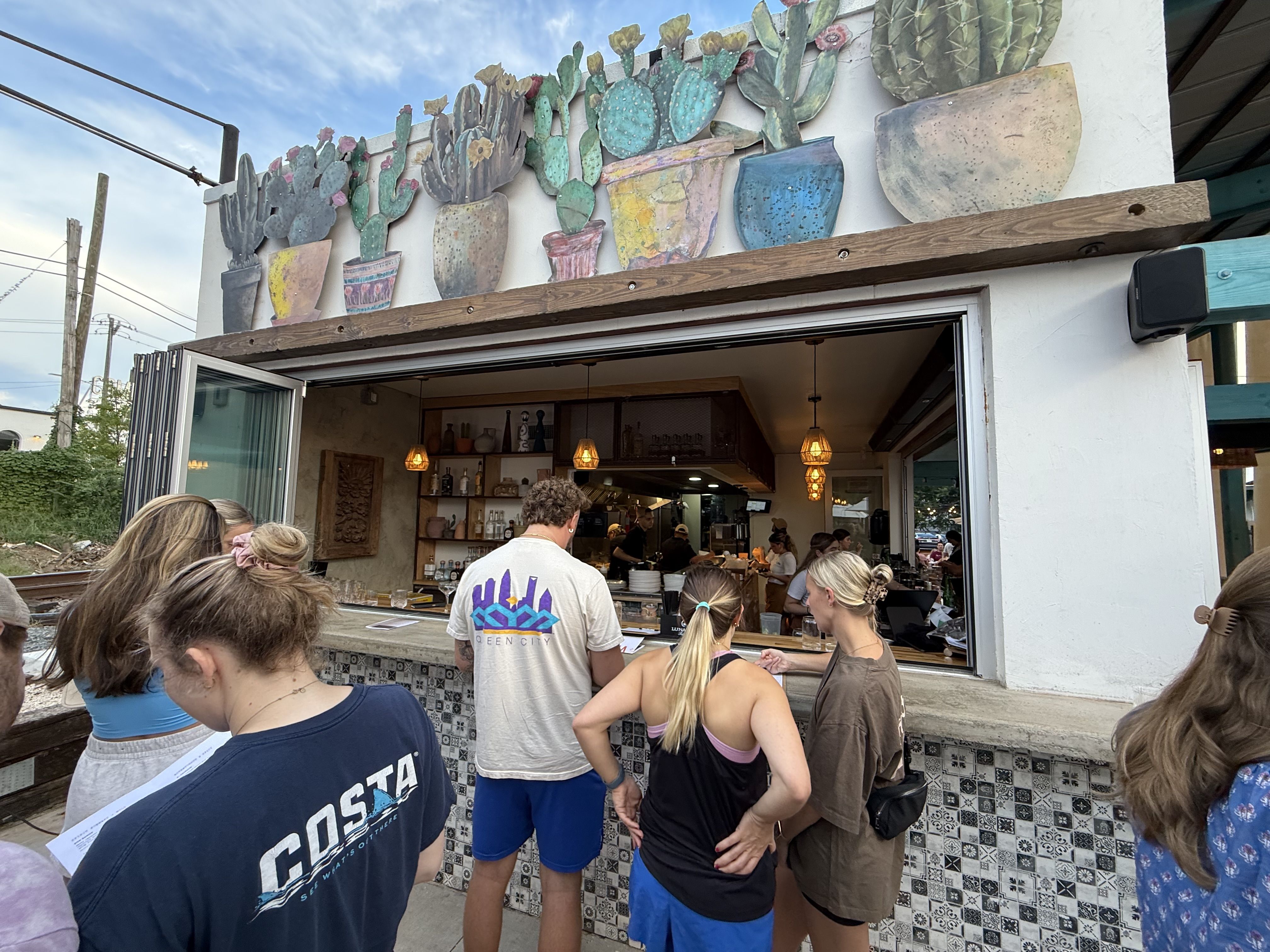 Open-front cafe with a mural of colorful cacti in pots along the roofline. Inside, staff serve customers at a counter while a crowd waits outside; blue sky and power lines above.