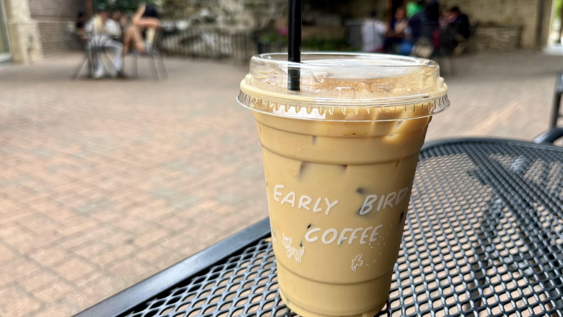 A disposable cup filled with a light-colored coffee sits on an iron bistro table. 