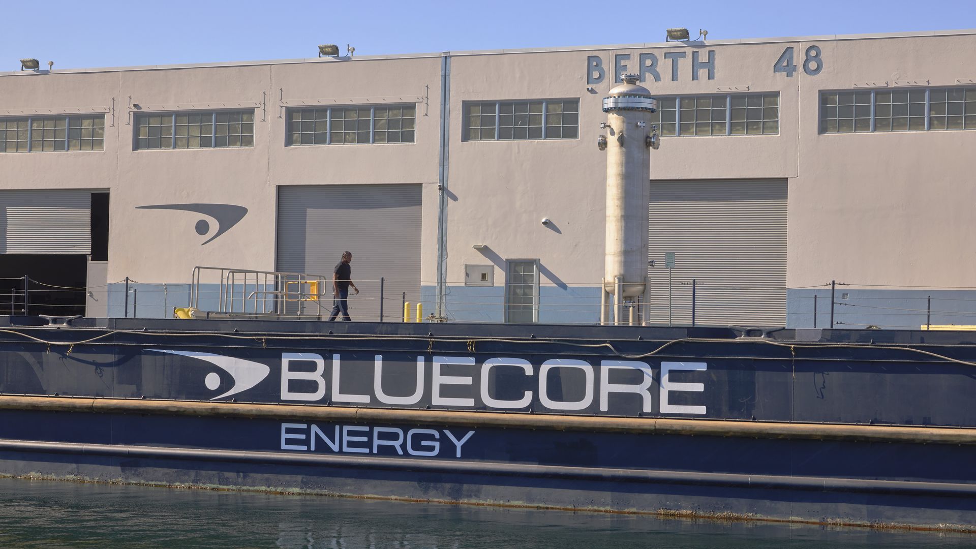 Beige port building labeled 'BERTH 48' with a tall vent pipe, a man walking on the dock, and a large hull reading 'BLUECORE ENERGY' beneath a clear blue sky.