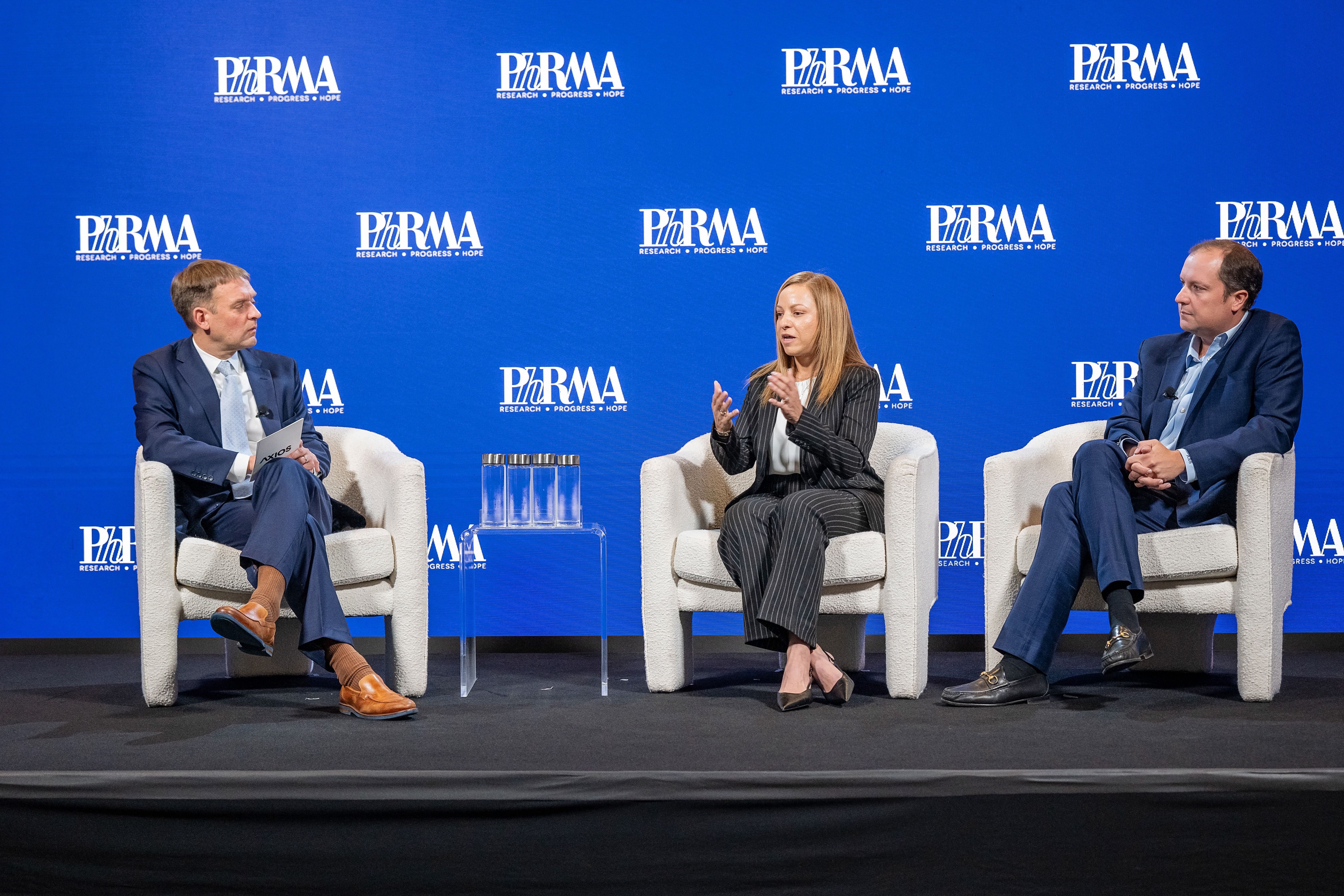 Three panelists in suits sit in white chairs on a blue PhRMA stage; a clear glass table with glasses sits in front of the middle speaker as she speaks.