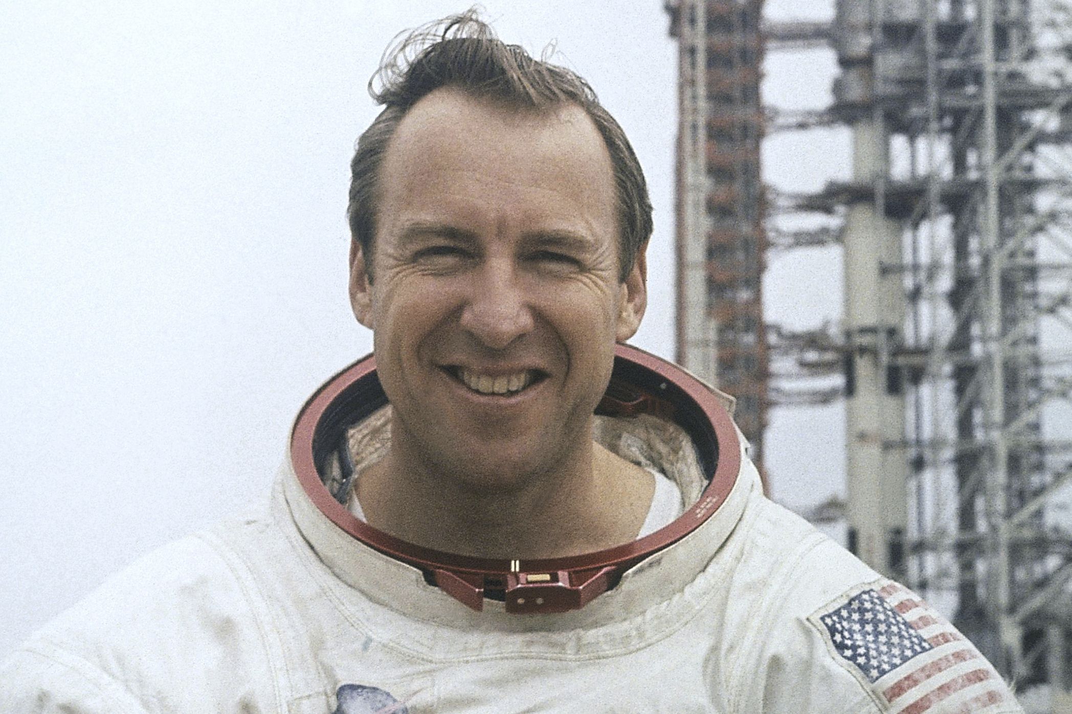 Man wearing a white astronaut suit with a red collar and U.S. flag patch, standing in front of a metal launch structure.