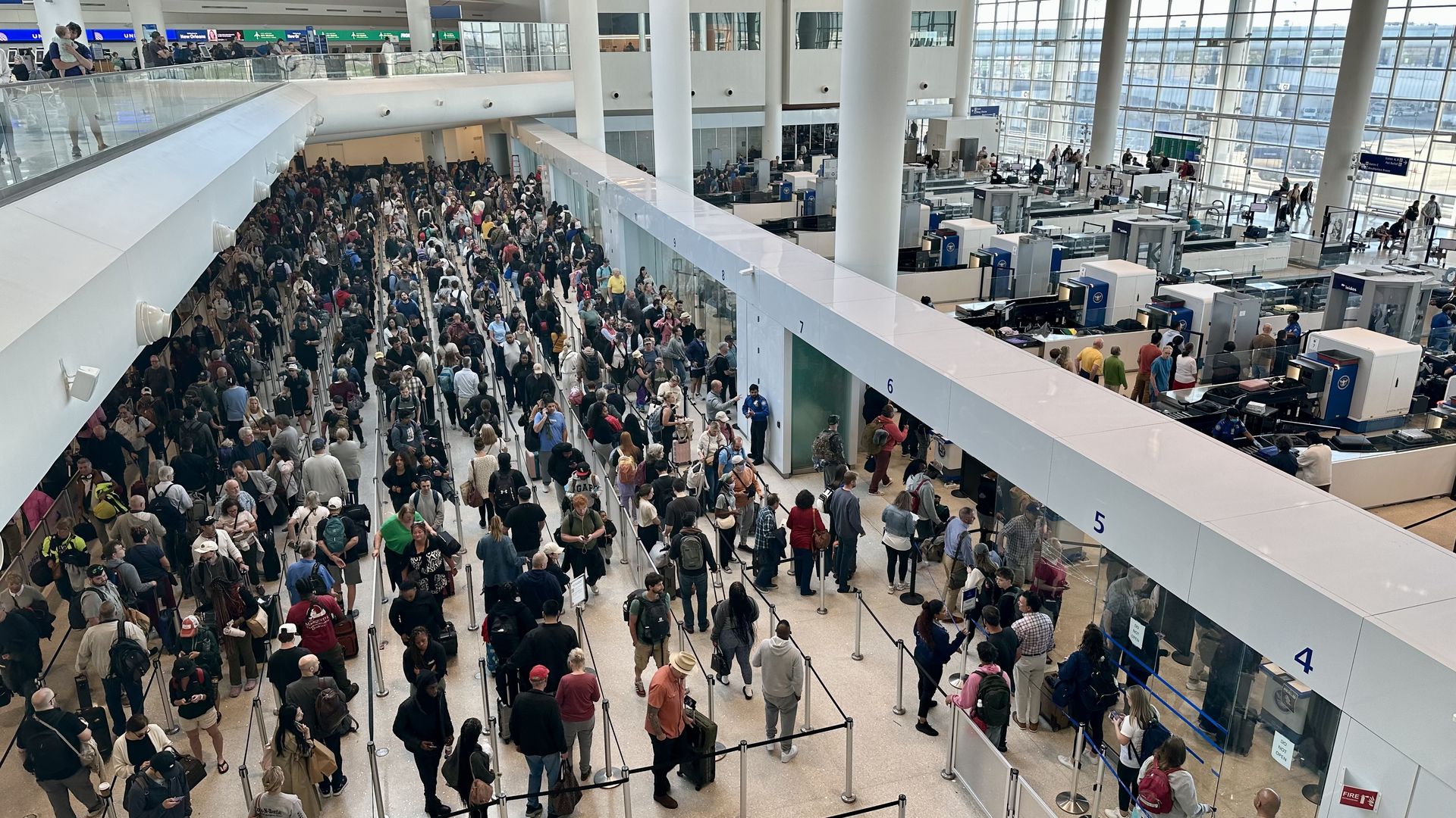 Busy airport security area with long lines of travelers and rolling luggage behind crowd-control stanchions; check-in desks on the right and glass walls in the background.