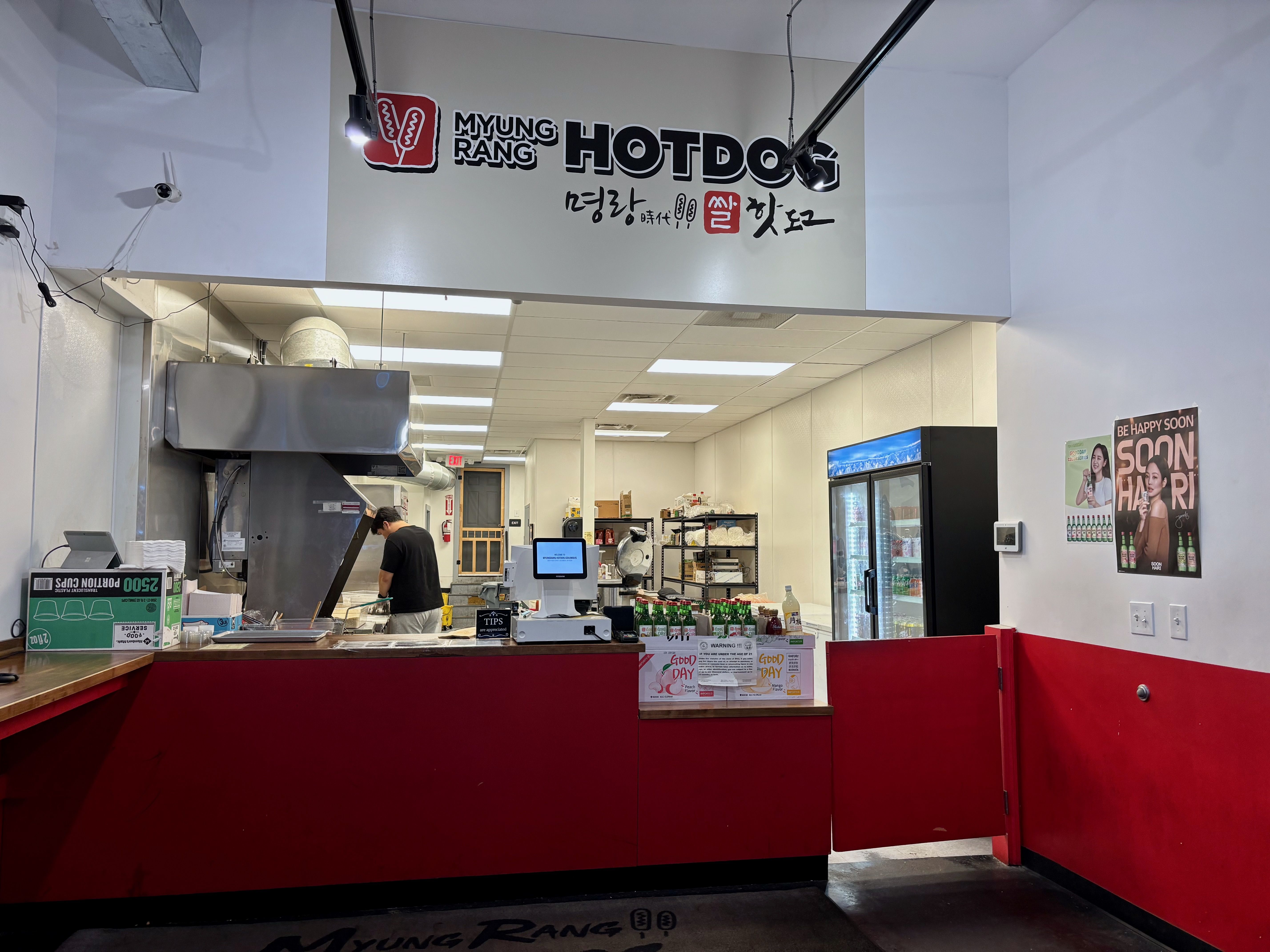 Interior of Myungrang Hotdog shop with red counter, a man working in kitchen, cooler with drinks, posters on white walls, and a sign with logo and Korean text above.