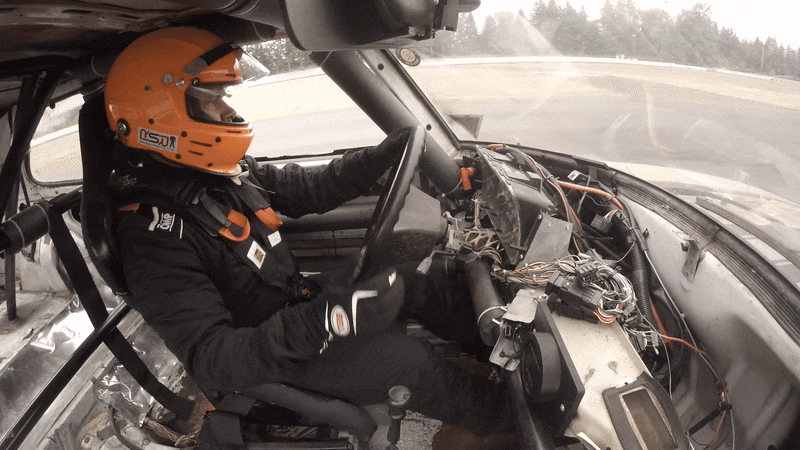 The inside of a stripped down race car shows the driver in a helmet glancing over as smoke comes out of the dashboard.