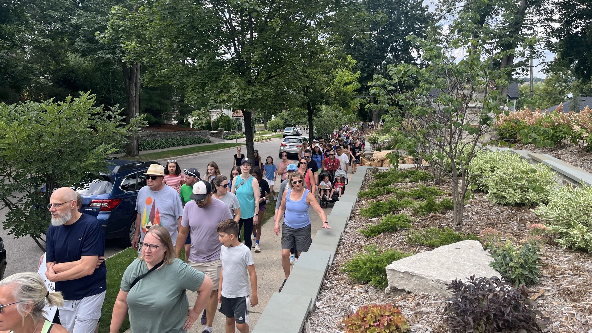Large group of people walking along a tree-lined sidewalk under a partly cloudy sky, next to a landscaped garden and parked cars on a residential street.