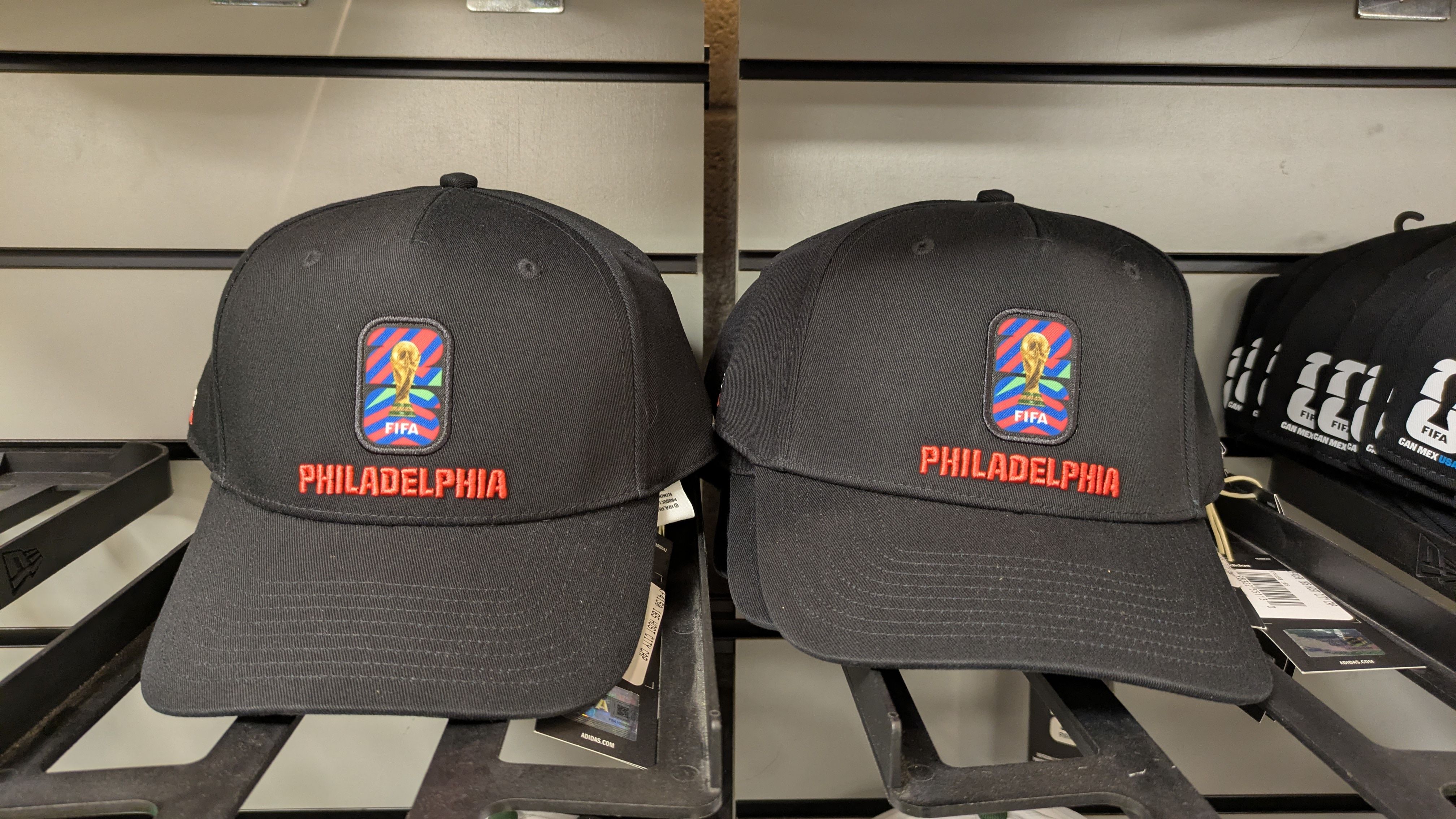Two black Philadelphia baseball caps with red embroidered "PHILADELPHIA" and a FIFA badge patch on the front, displayed on a store shelf.