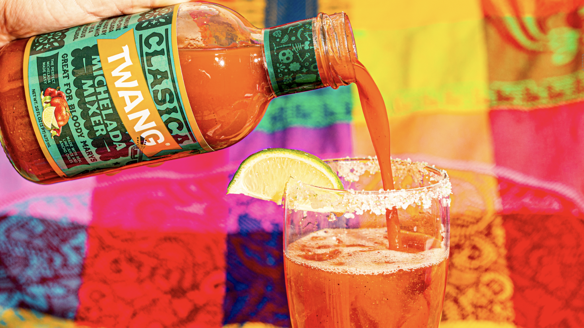Hand pouring orange-red Twang Classic Michelada Mixer into a salt-rimmed glass with lime wedge, set against a vibrant multicolored background.