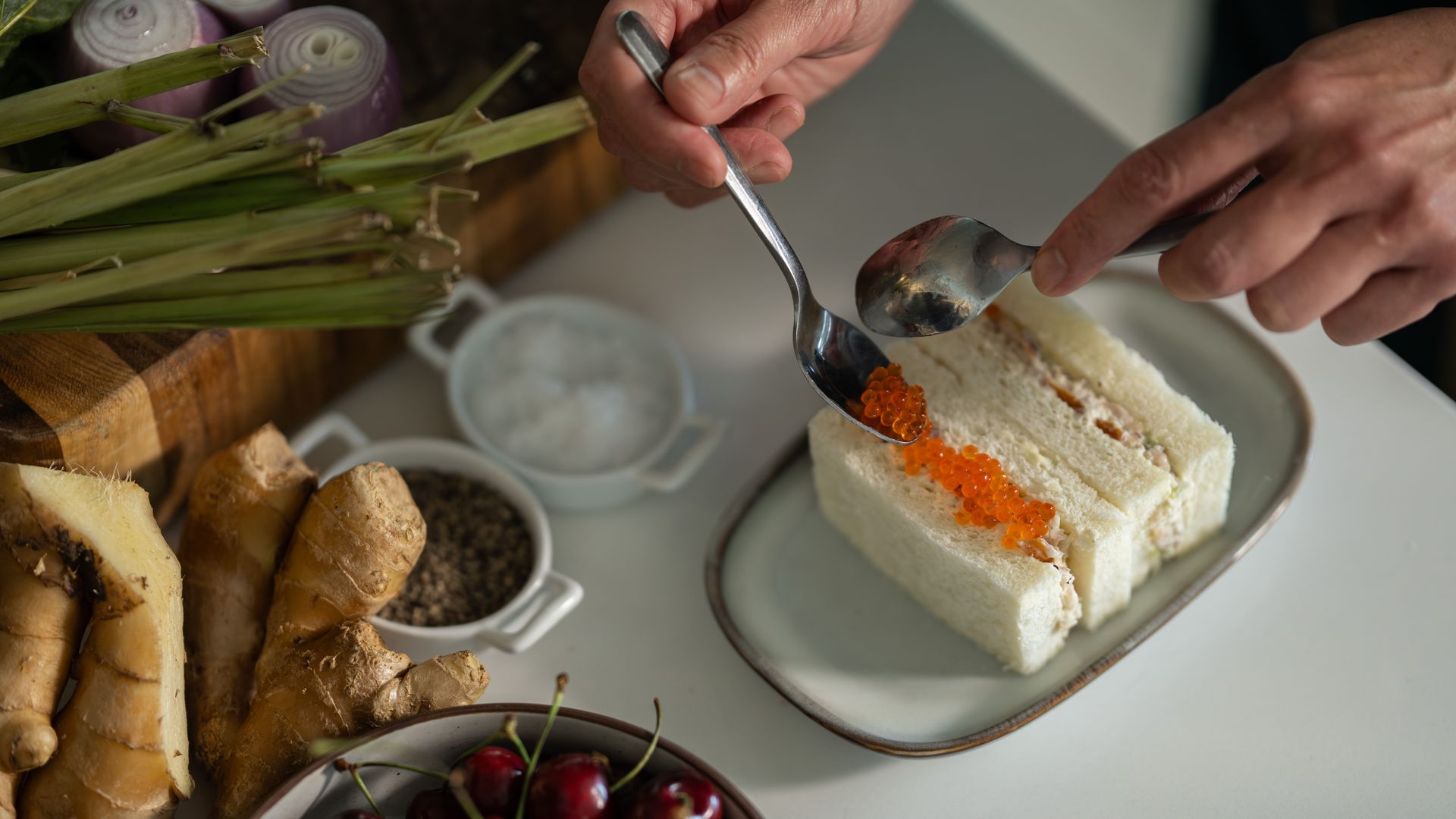 Close-up of a sandwich plate as a hand adds bright orange salmon roe with a spoon; background shows leeks, red onions, bread, and a bowl of cherries.