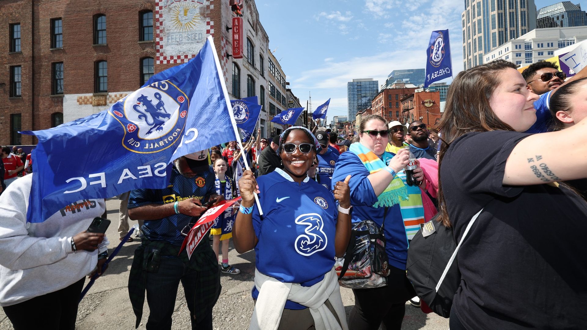 A woman wears a blue Chelsea Premier League jersey and carries a Chelsea flag in a sea of people walking down a city street.