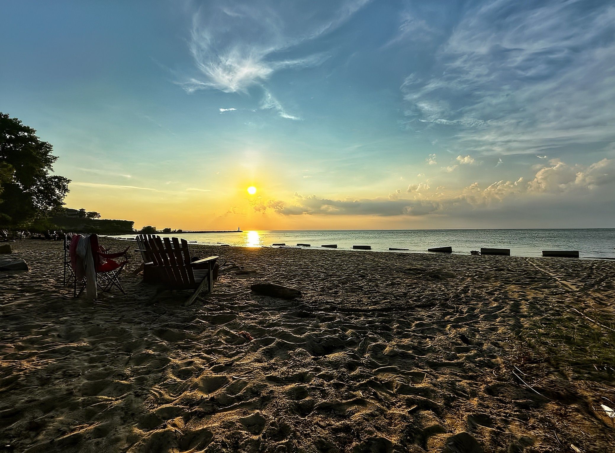 Sunset over a sandy beach with calm water and scattered clouds. Two wooden chairs and a red folding chair are on the beach, with trees on the left side.