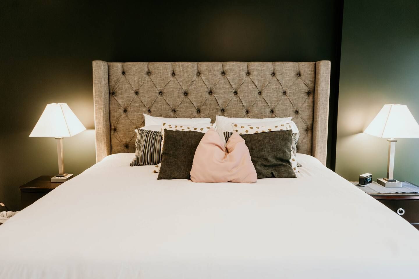 King-sized bed with a tufted beige headboard, white bedding, and a mix of striped, patterned, dark gray, and pink pillows, flanked by two lit table lamps on dark nightstands.