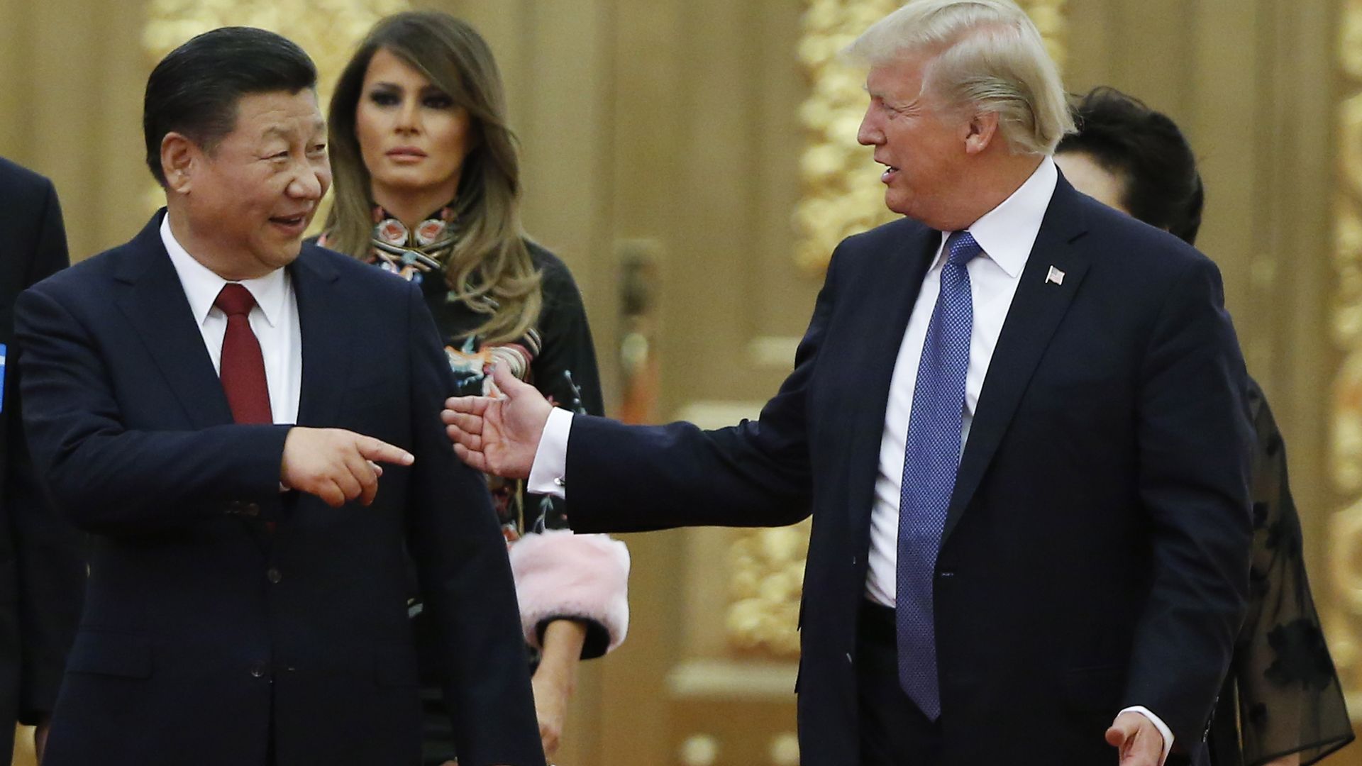 Trump and Xi JingPin gesture toward one another. Melania Trump is in the background. 
