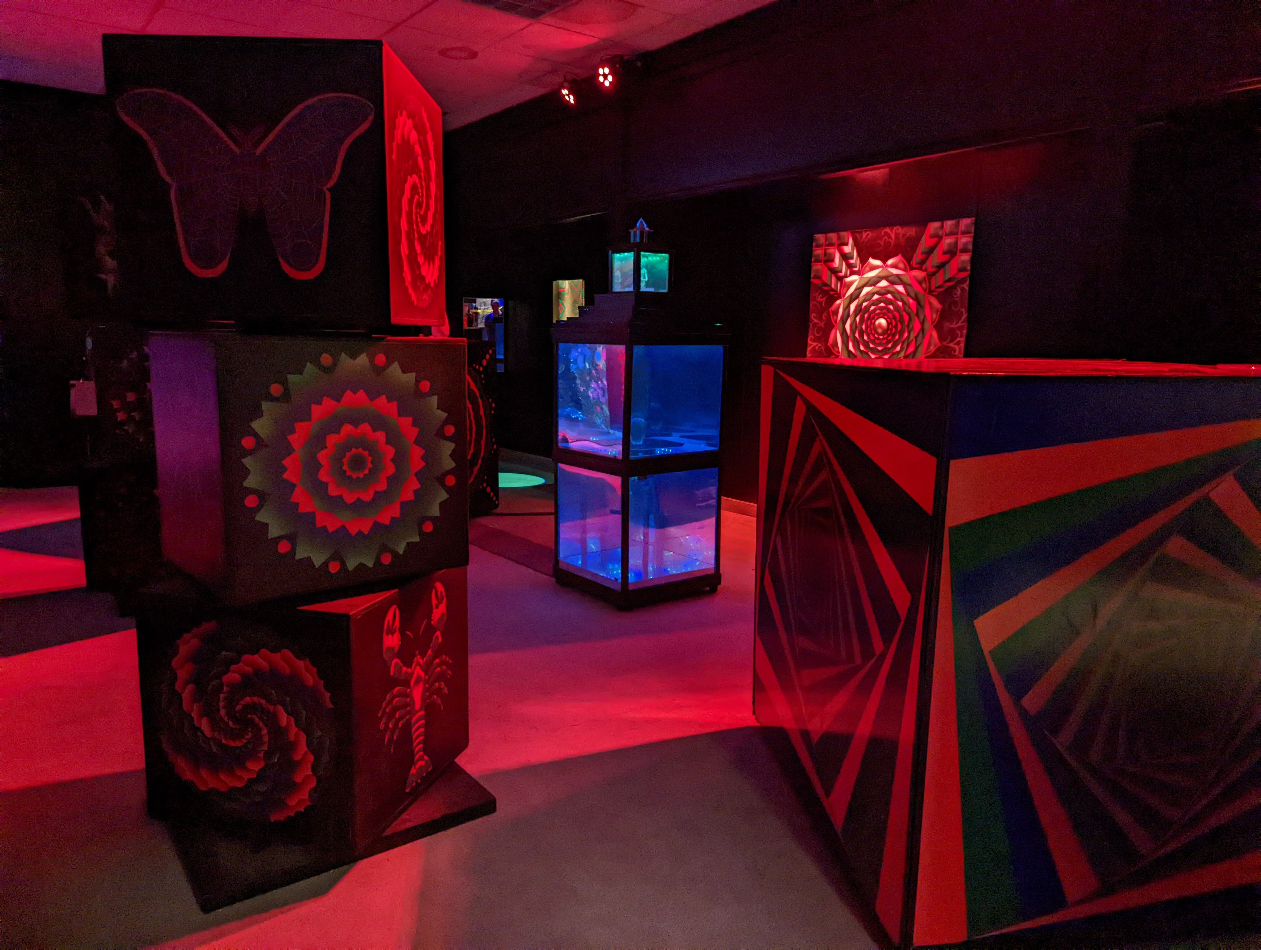 Geometric shapes are painted on columns of boxes under red lights in an art gallery.