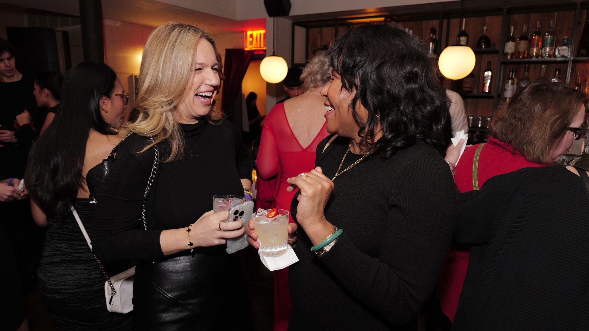 Two women wearing black tops laugh and hold cocktails at a lively bar party with a wooden shelf of liquor bottles and warm pendant lights in the background.