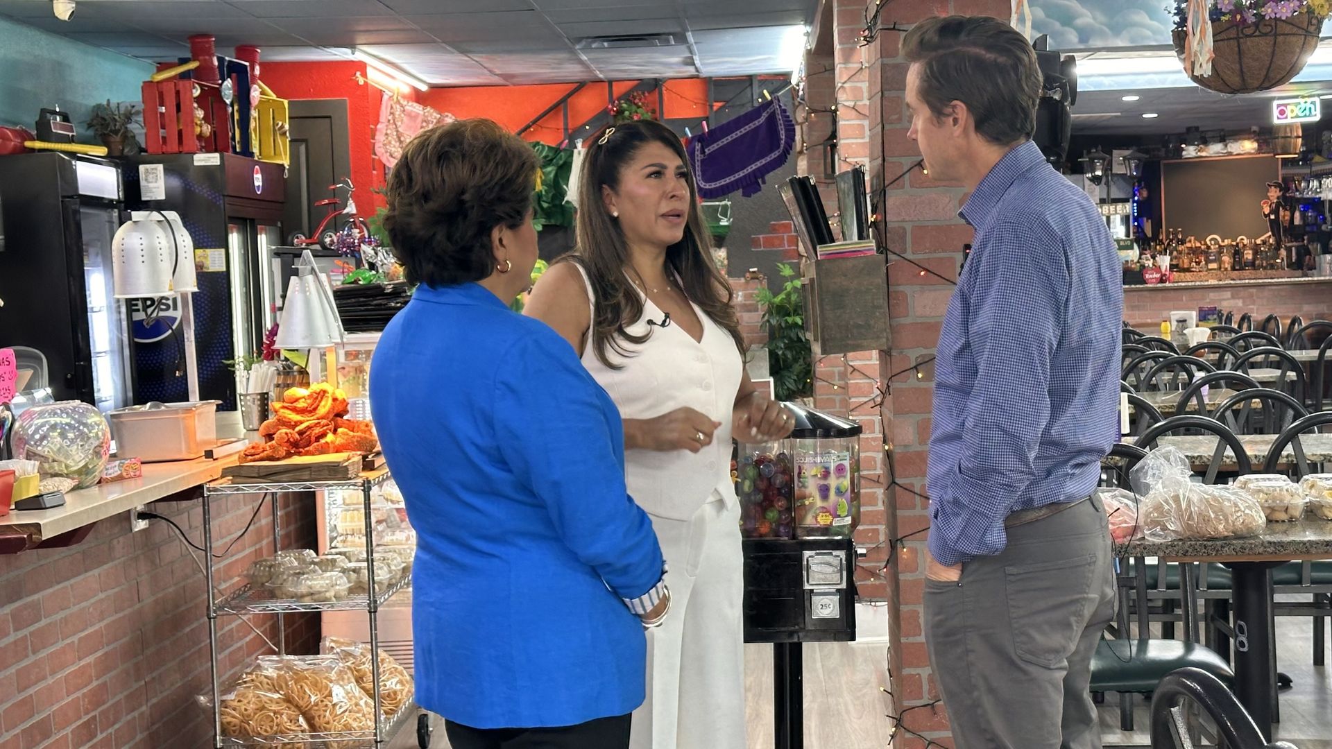 A photo of three people chatting in front of a cafe counter: a woman in blue, a woman in white, and a man in a blue checkered shirt. Pastries and drinks line the counter; brick walls and a bar are in the backdrop.