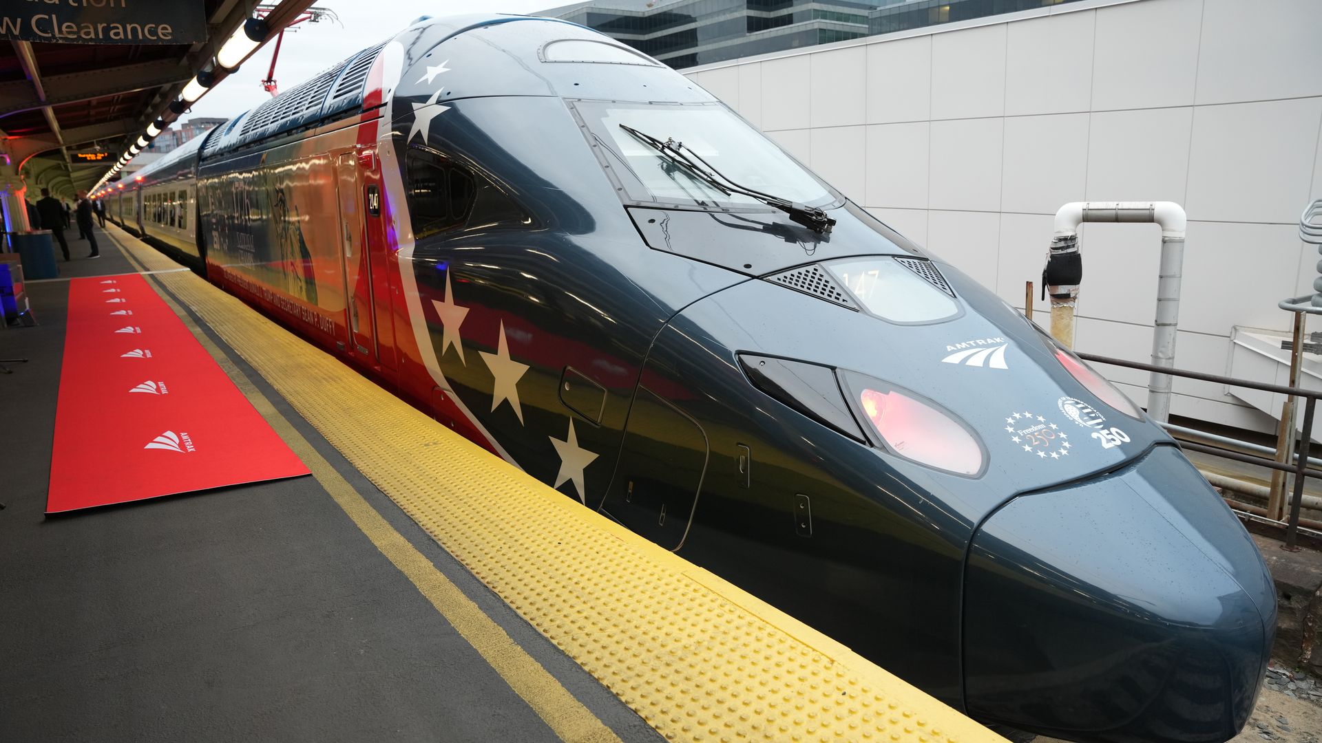 Front-right view of a gray Amtrak high-speed train at a platform, white star decals along its side, yellow tactile paving and a red safety mat, with city buildings in the background.