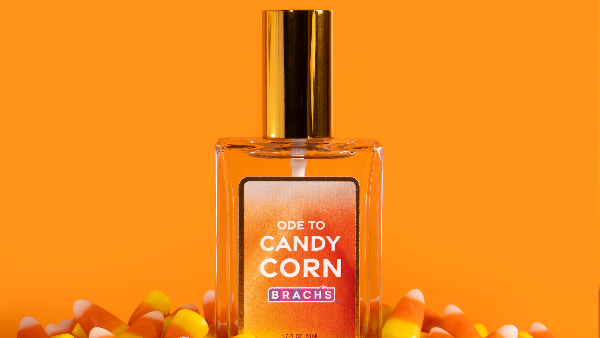 Close-up of a perfume bottle with a shiny gold cap on an orange background, label reads "ODE TO CANDY CORN" surrounded by colorful candy corn pieces.