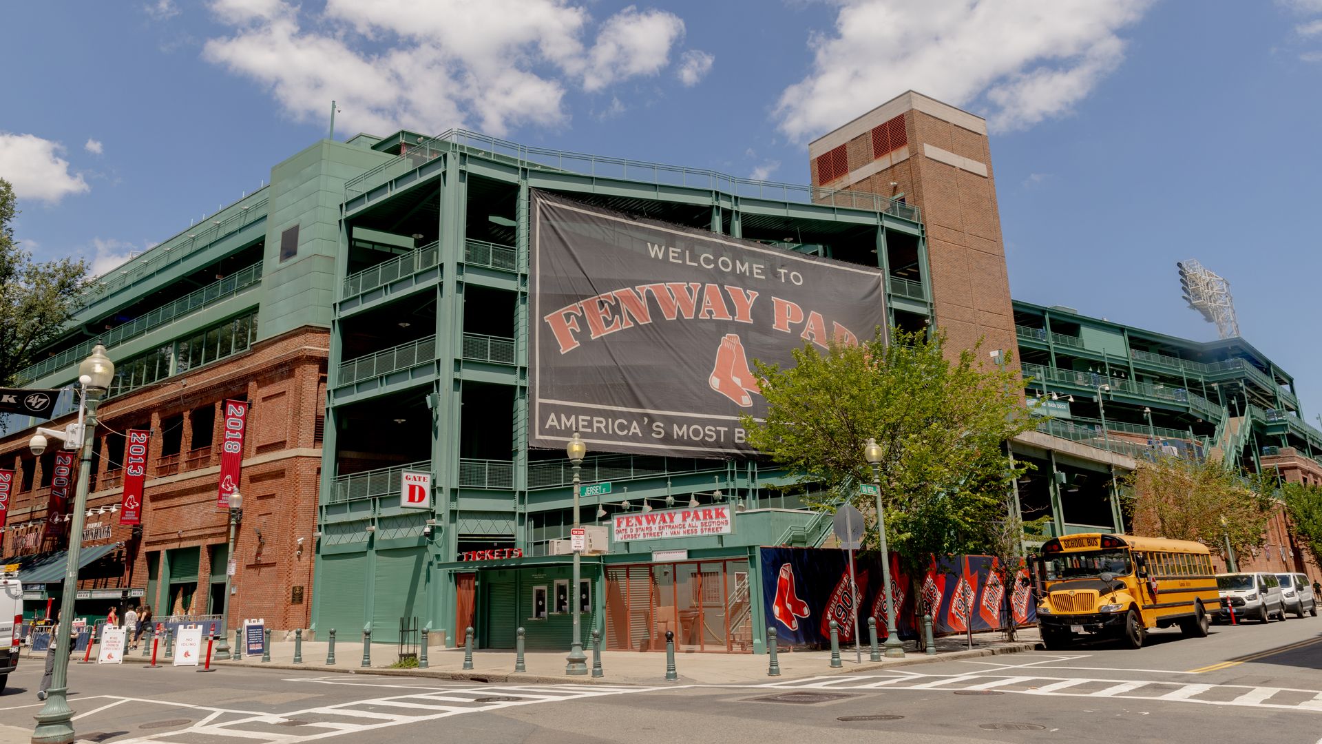 Exterior view of Fenway Park baseball stadium in Boston with a large welcome banner, red brick and green steel structure, under a blue sky with scattered clouds and a yellow school bus nearby.