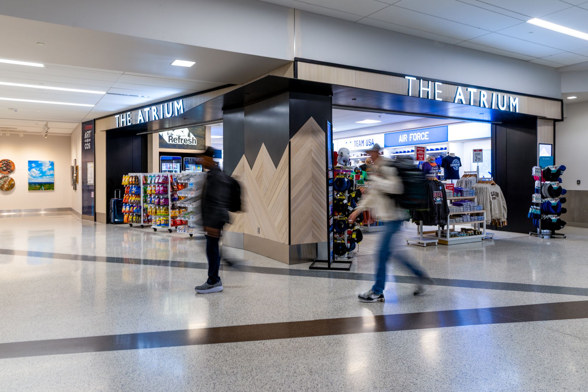 Inside a mall atrium, THE ATRIUM storefront in bright letters. Wood-framed entrance, mountain pillar; blurred shoppers pass as snack racks and a blue AIR FORCE shop are visible.