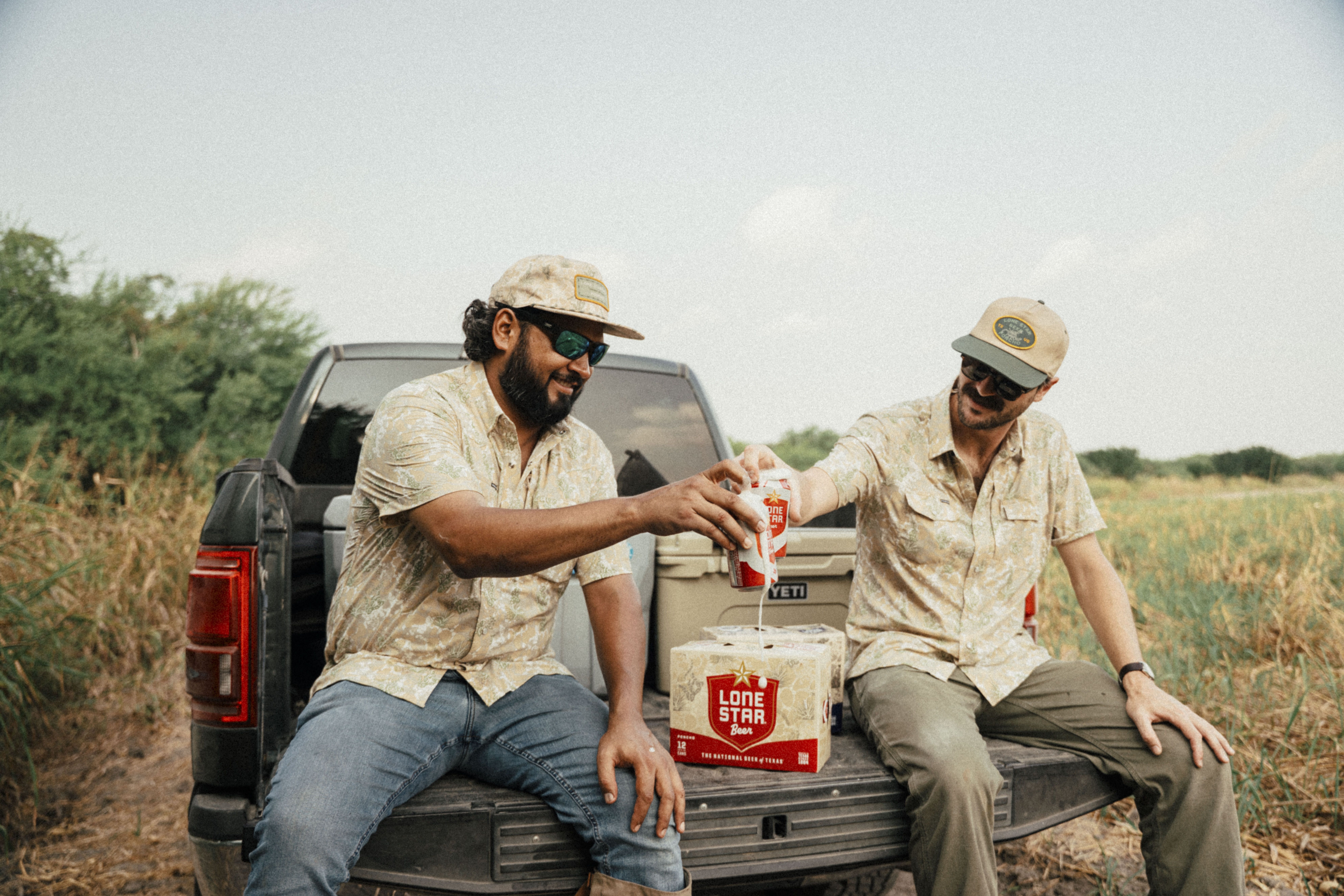 Two men wearing patterned shirts, sunglasses, and caps sitting on a truck tailgate in a field, sharing and clinking cans of Lone Star Beer on a sunny day.