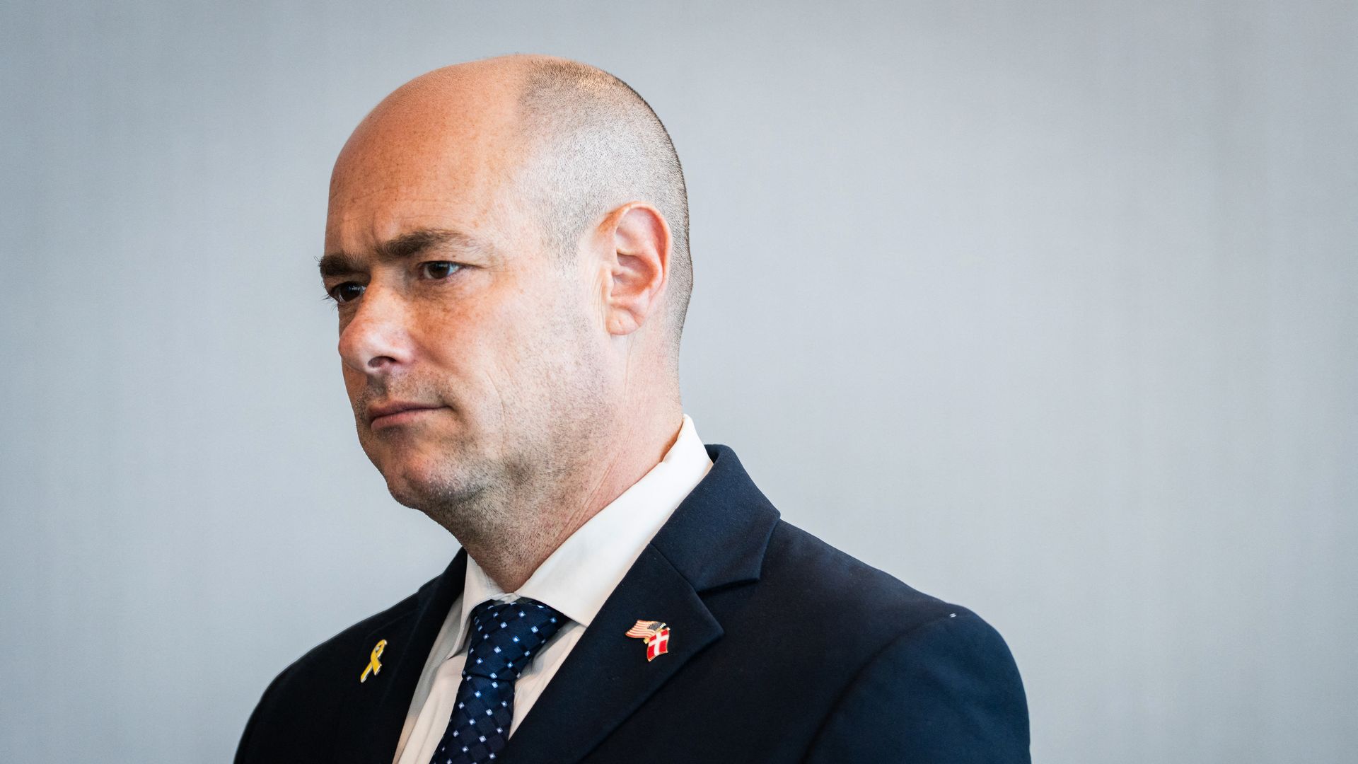 Man wearing a navy suit, white shirt, and blue polka dot tie, with yellow and flag pins on his lapel, against a plain light gray background.