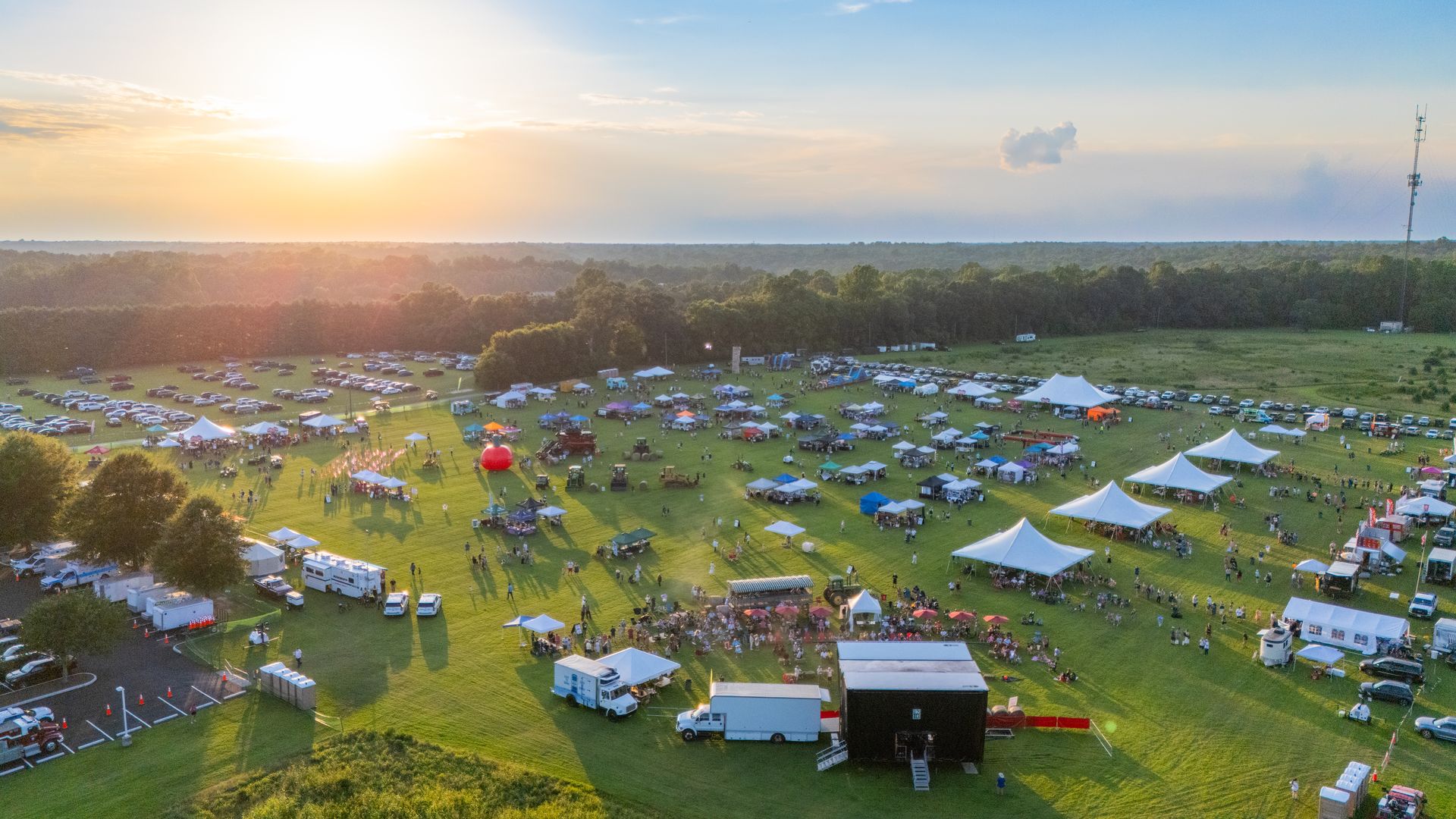 An aerial view of a large outdoor festival on a grassy field at sunset. Dozens of white tents and colorful canopies dot the area, with parked cars along the edges and a tall radio tower on the right.