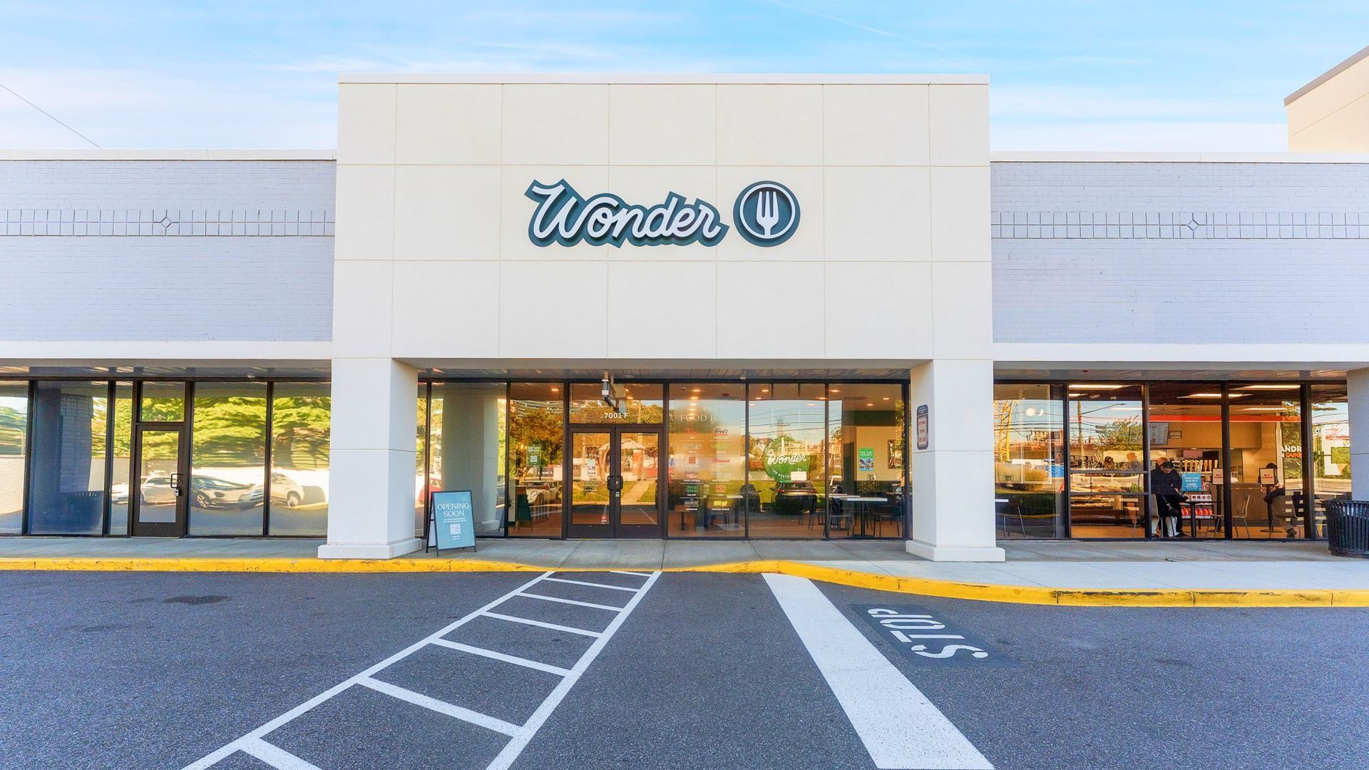 Front view of a food hall named Wonder with large glass windows and doors, a white facade, a blue sign, and a parking lot with white lines and a stop marking.