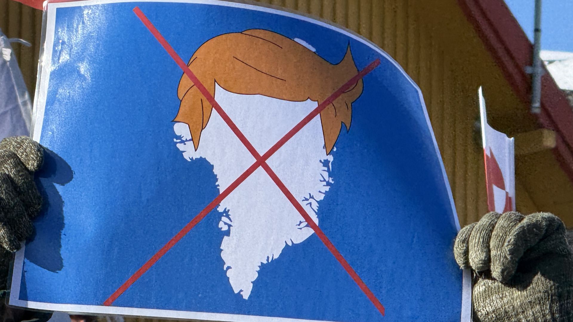 Person wearing gray gloves holds a blue sign with a white map of Greenland crossed out with red lines and a cartoon orange hair silhouette above the map on a sunny day.