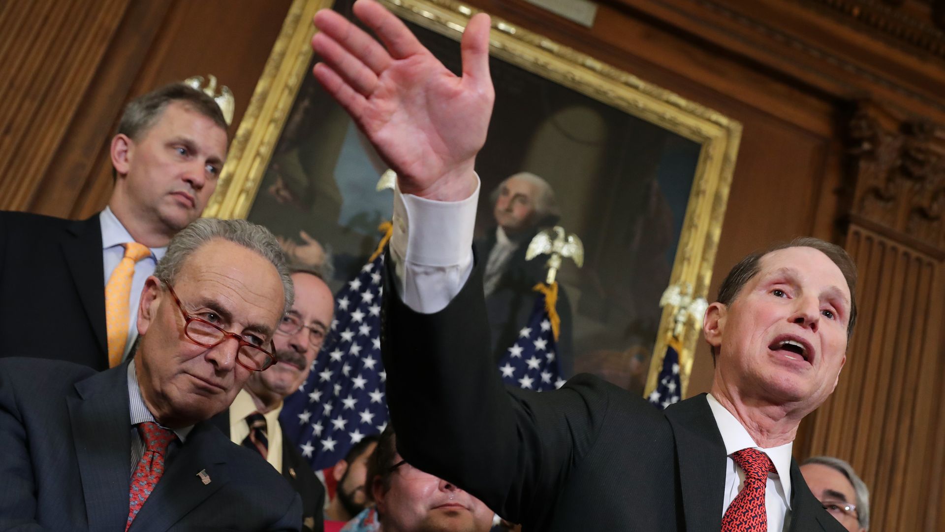 Chuck Schumer watches as Ron Wyden raises his hand while giving a speech. 