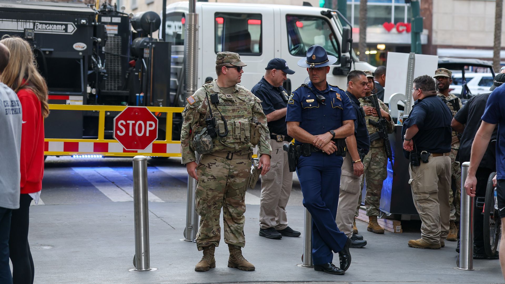Group of uniformed military and police personnel standing near a stop barrier on a city street with a white truck and building in the background.