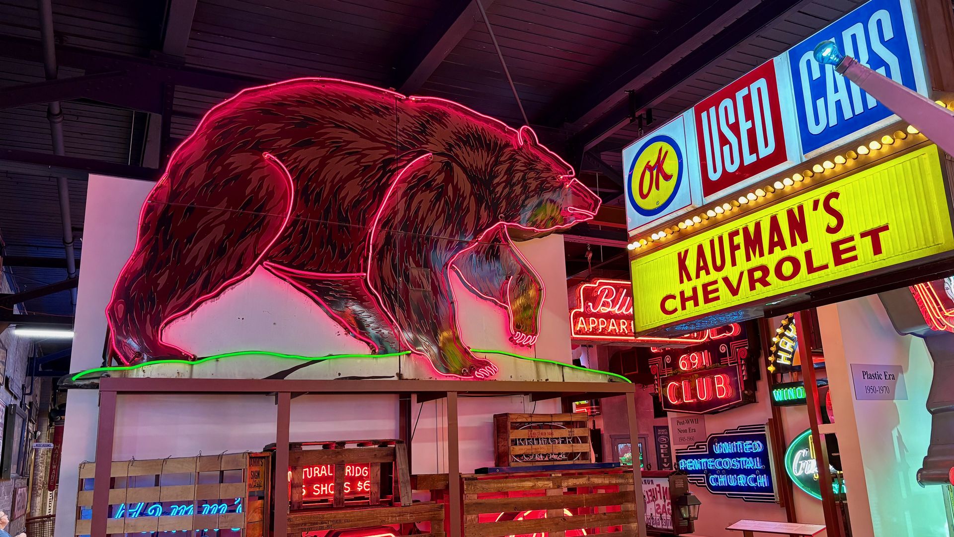 Indoor retro showroom featuring a giant pink neon bear outline, blue "USED CARS" sign, and yellow "KAUFMAN'S CHEVROLET" sign amid colorful neon lights and wooden railings.