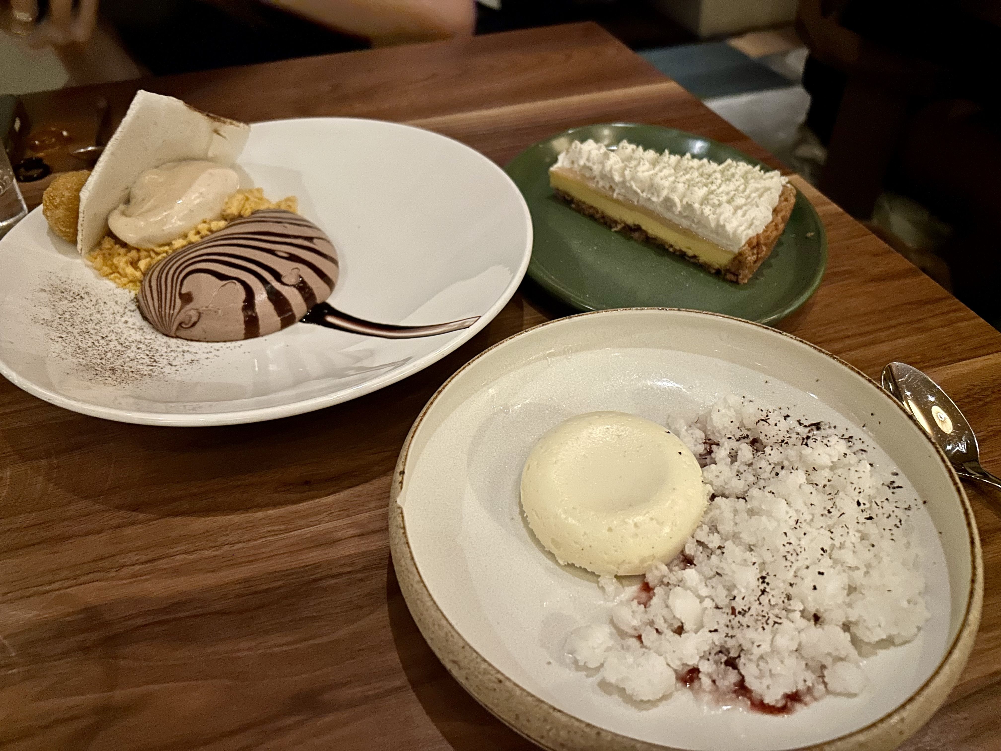 Three plated desserts on a wooden table: a swirl chocolate mousse with cream and crispy elements on a white plate, a whipped cream-topped pie slice on a green plate, and a panna cotta with crumbled ice and sauce on a beige plate.