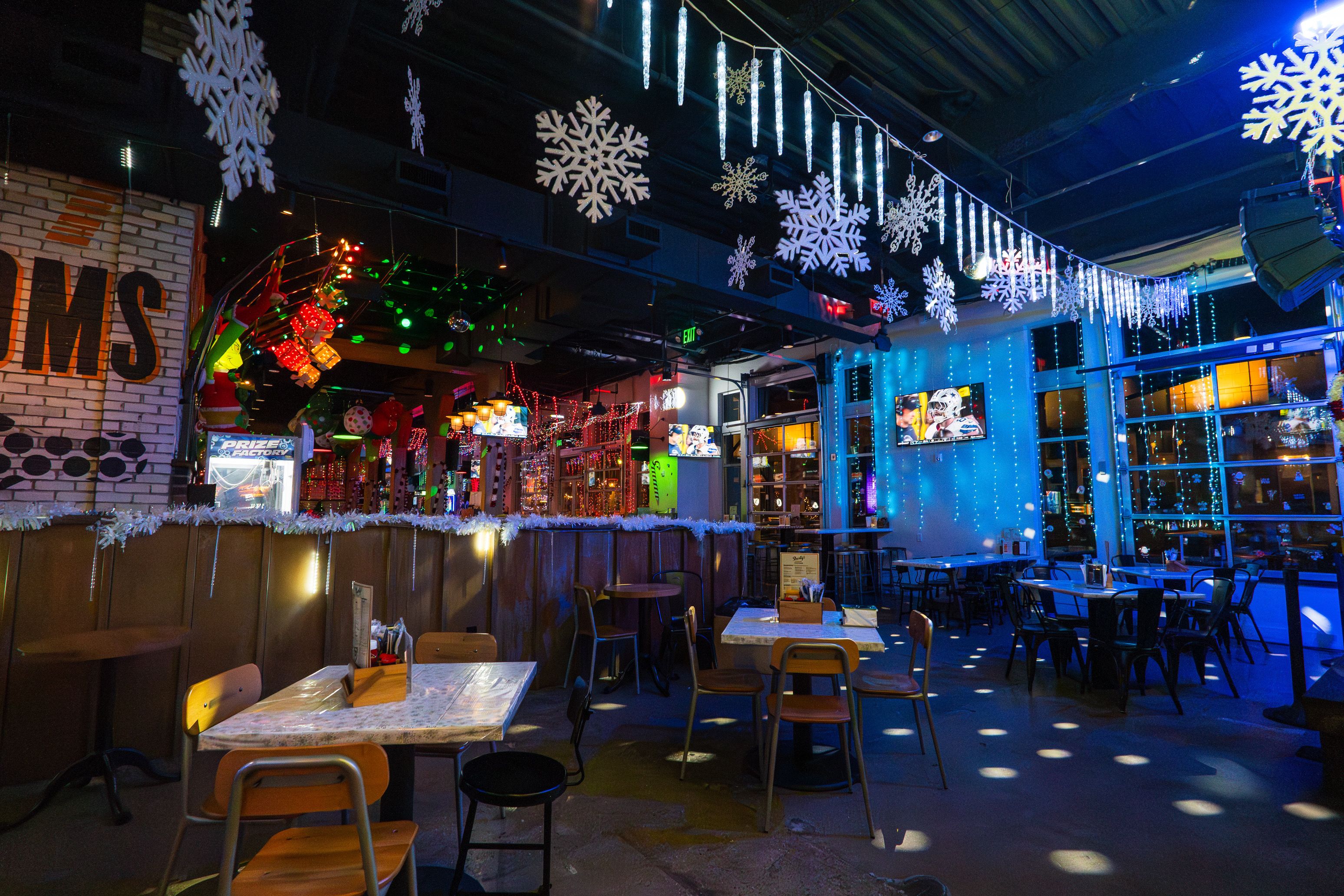 Indoor dining area decorated with hanging white snowflakes and icicle lights, blue wall lights, multiple tables and chairs, TV screens showing sports, and festive red and green string lights.