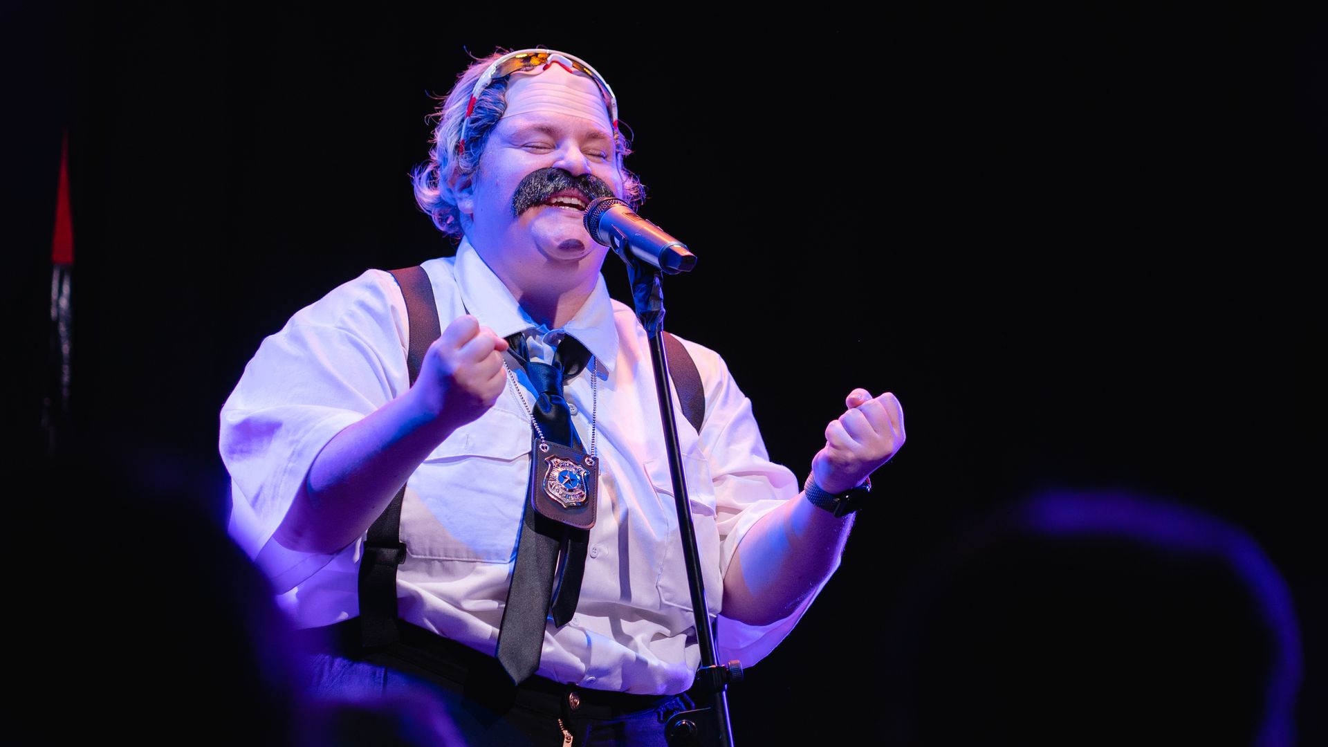 A woman dressed as a balding police officer with a mustache smiles and speaks in a microphone