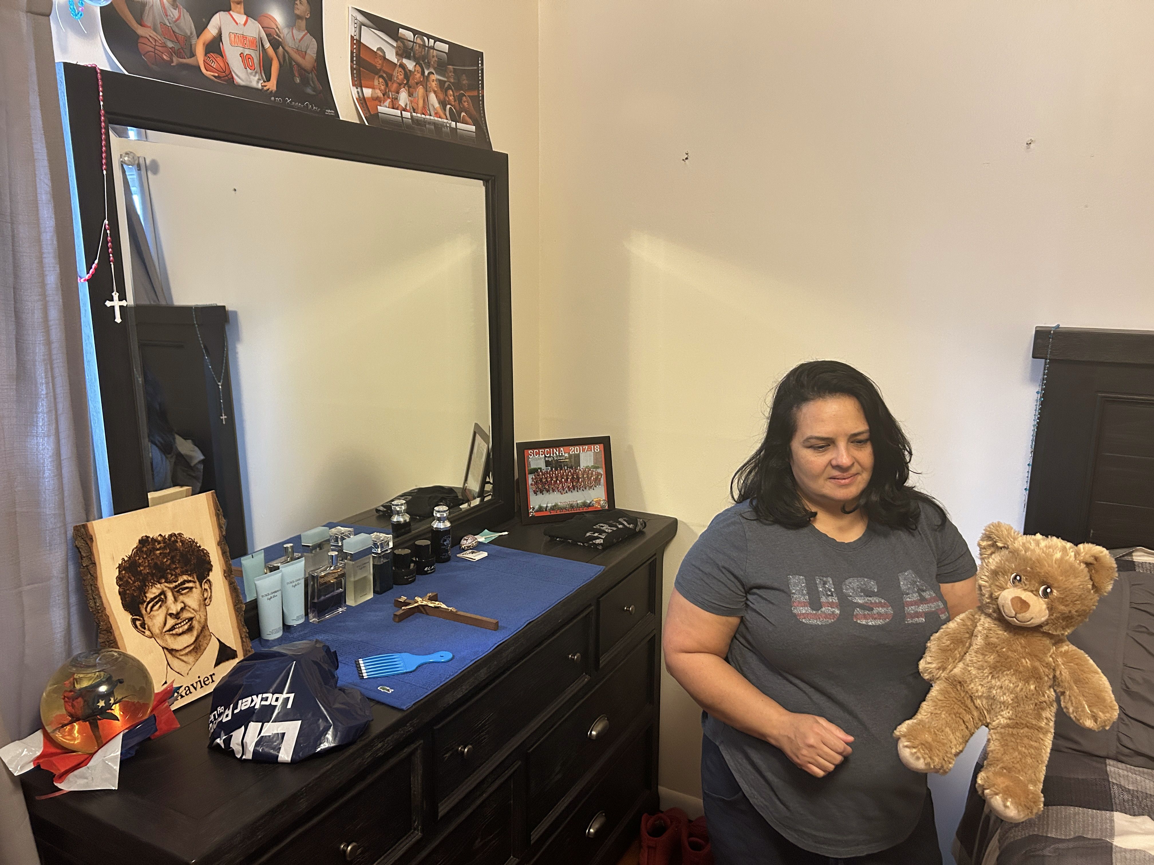 Woman wearing a gray "USA" t-shirt holding a brown teddy bear in a bedroom with a dark dresser, a large mirror, and various items including perfumes, a crucifix, and photos on the dresser.