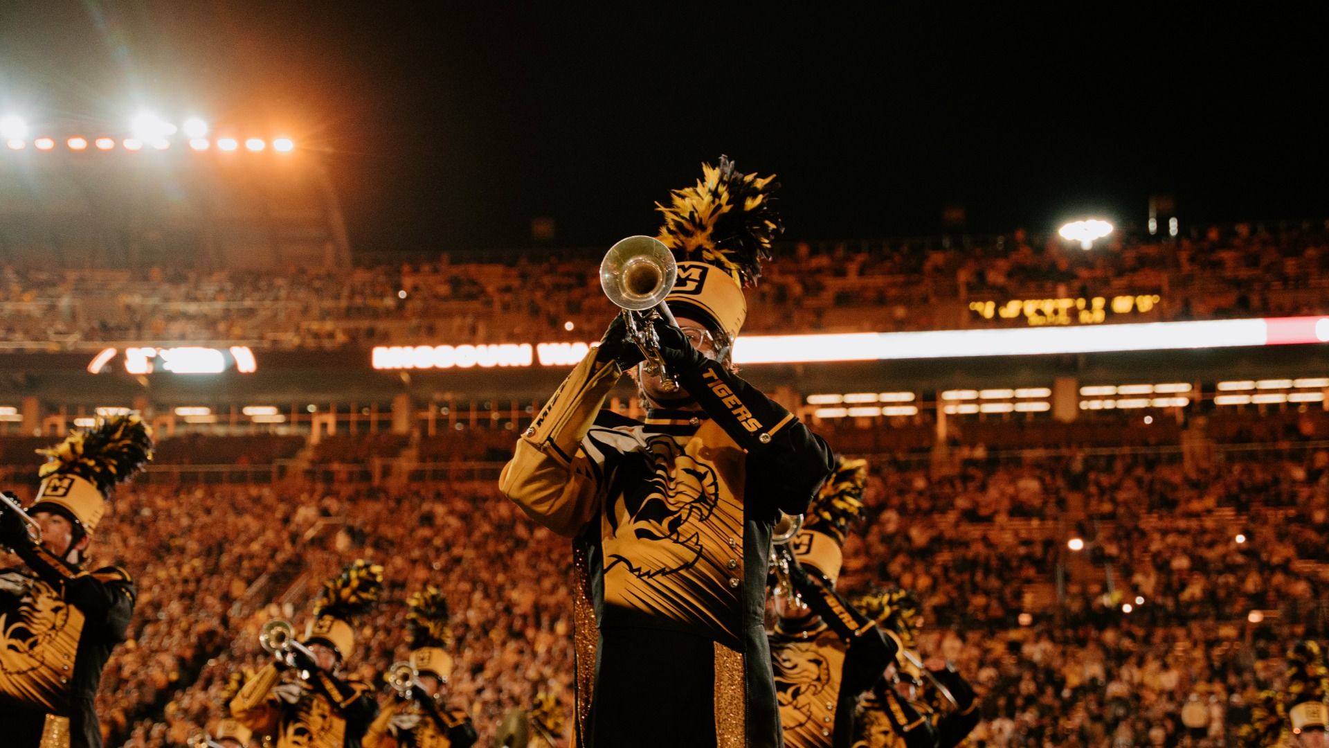 A marching band performs in a crowded stadium at night. A trombone player in a yellow-and-black uniform with a feathered plume raises a brass instrument toward the crowd while bright stadium lights.