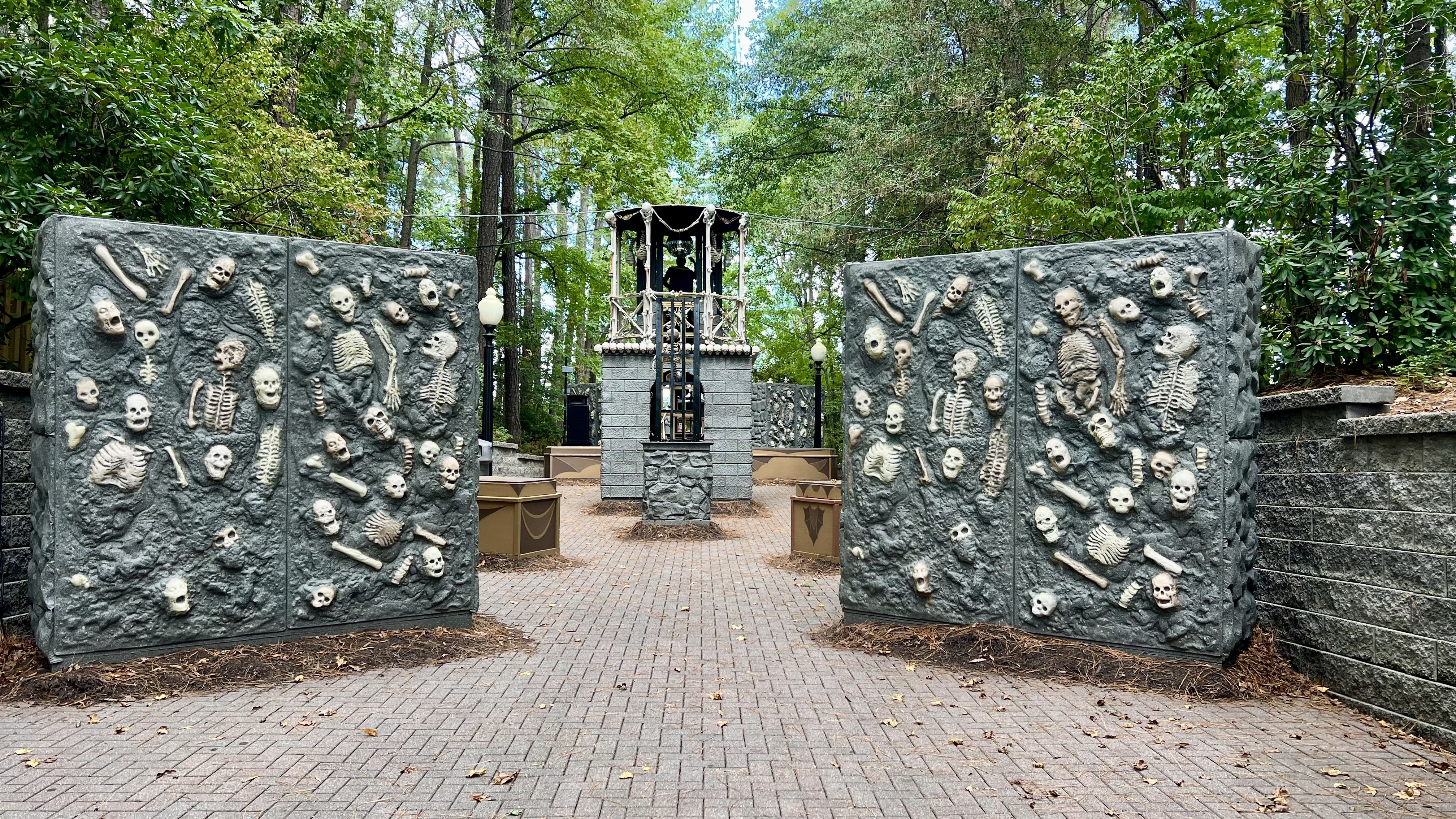 Outdoor scene featuring two large stone walls embedded with skull and bone sculptures, flanking a brick pathway leading to a stone well with a skeleton figure inside, surrounded by green trees.