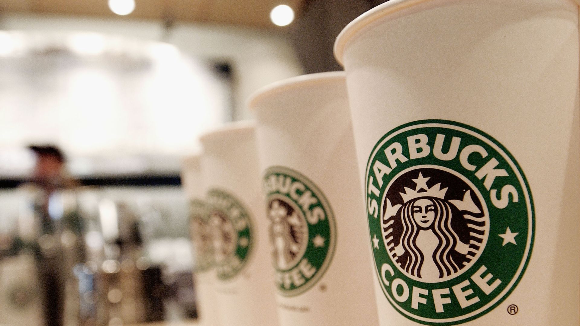 Four white Starbucks coffee cups in a row on a wooden counter, each displaying the green and black Starbucks logo, with a blurred coffee shop background.
