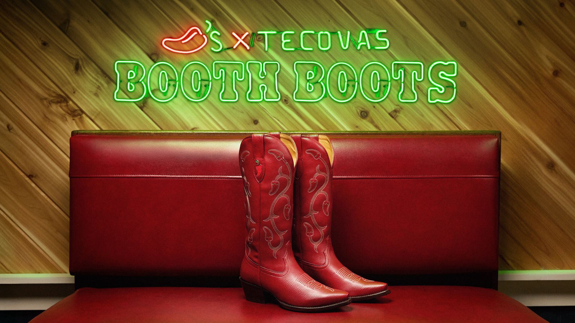 Pair of red leather cowboy boots with chili pepper designs, placed on a red leather bench in front of a wooden wall with green neon sign reading "Chili's X Tecovas BOOTH BOOTS".