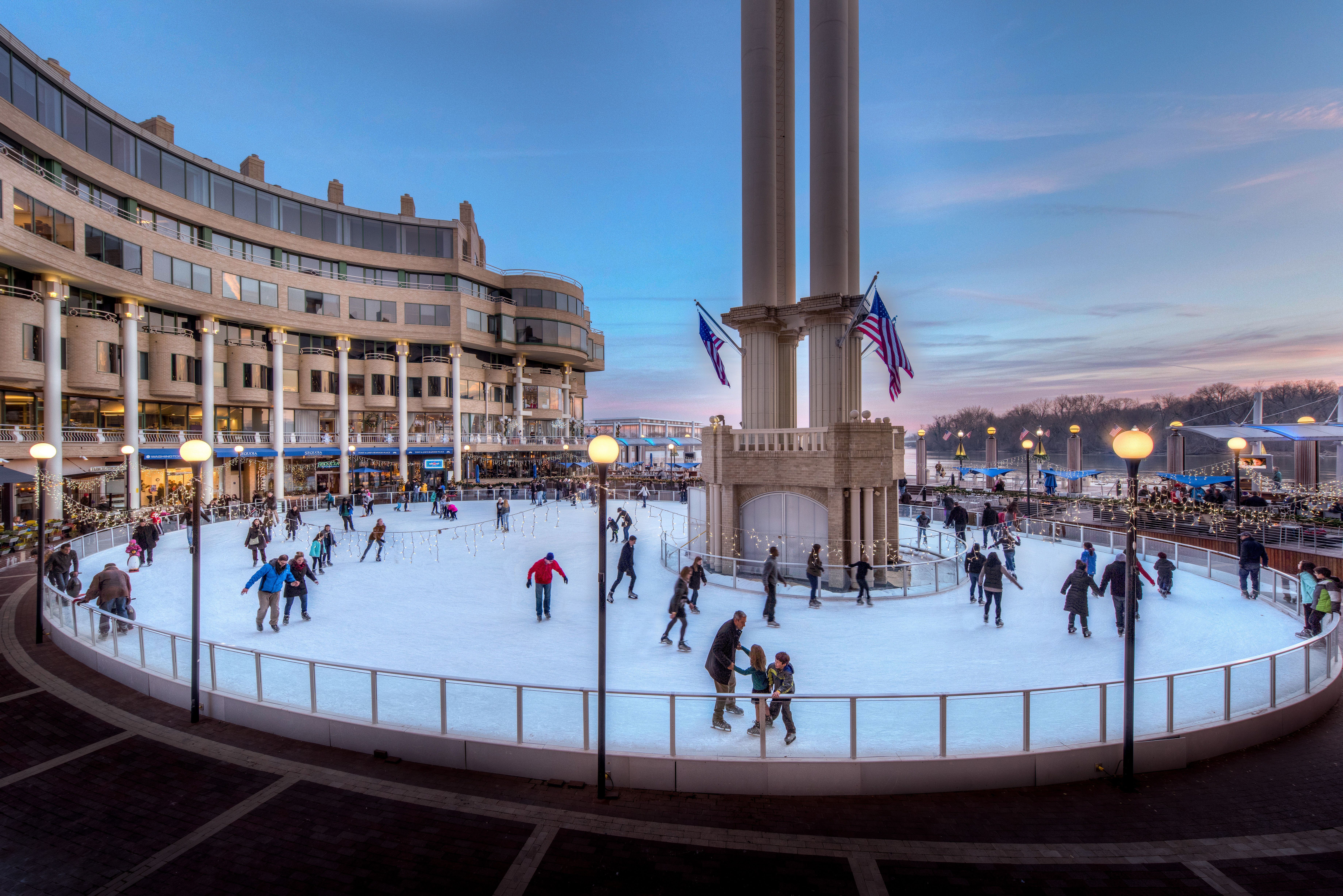 People ice skating on a large outdoor rink surrounded by a curved building and tall columns with American flags at sunset, with illuminated lampposts and festive lights.