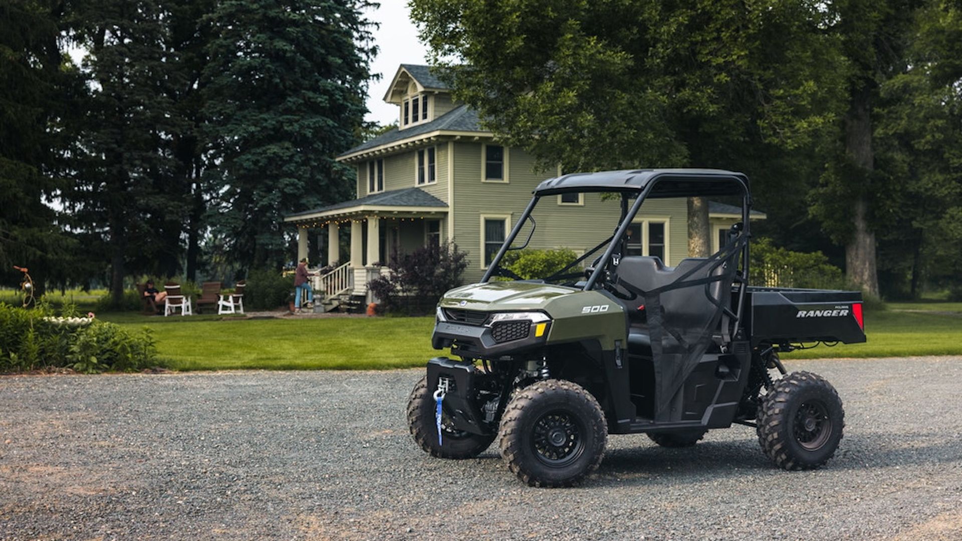 Green Polaris Ranger 500 utility vehicle parked on gravel in front of a two-story light green house with trees and lawn furniture nearby.
