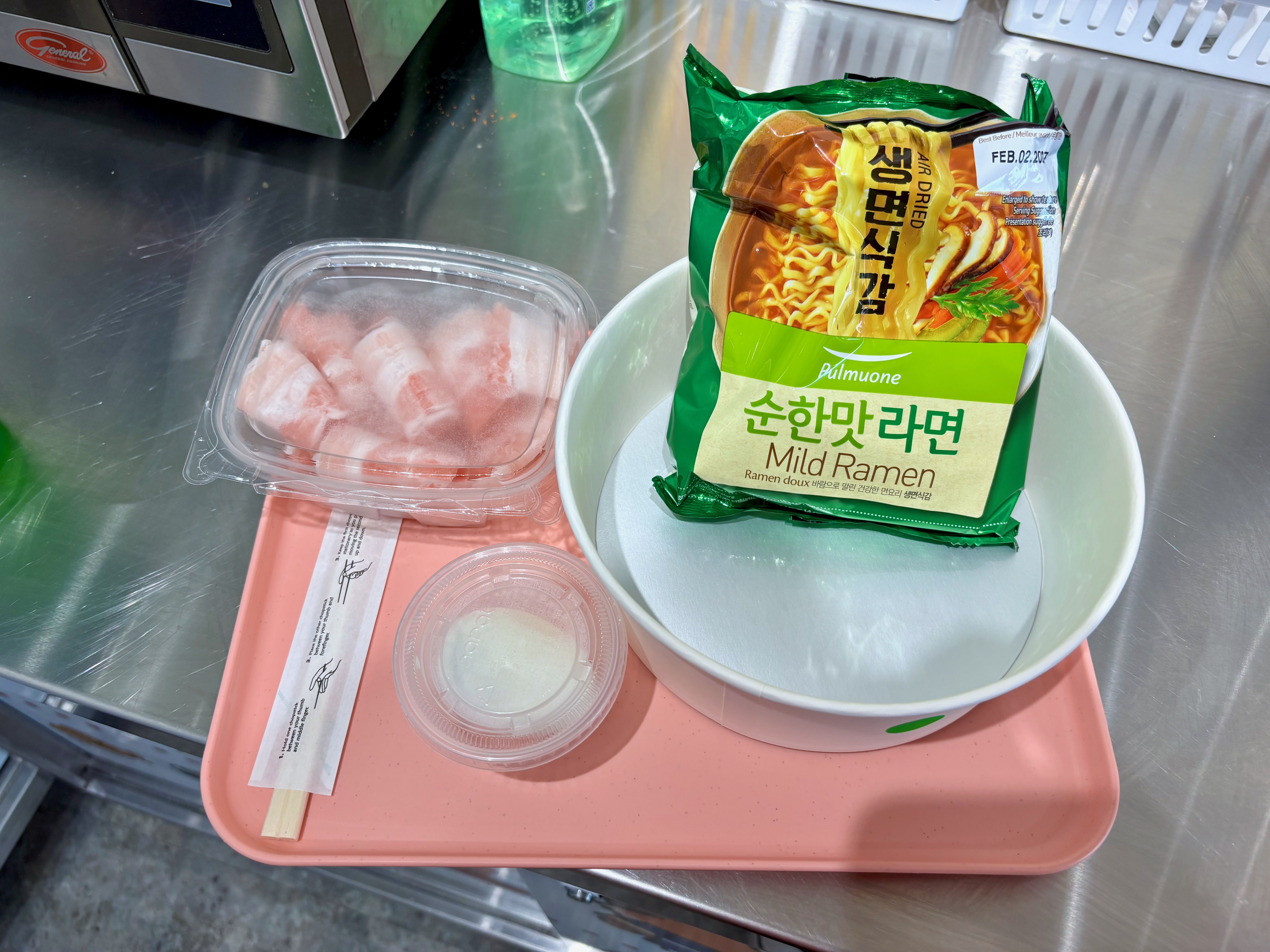 Stainless kitchen counter with a bright green bag of "Mild Ramen" noodles, a plastic container of pink raw meat on a pink tray, chopsticks, and a green detergent bottle in the background.